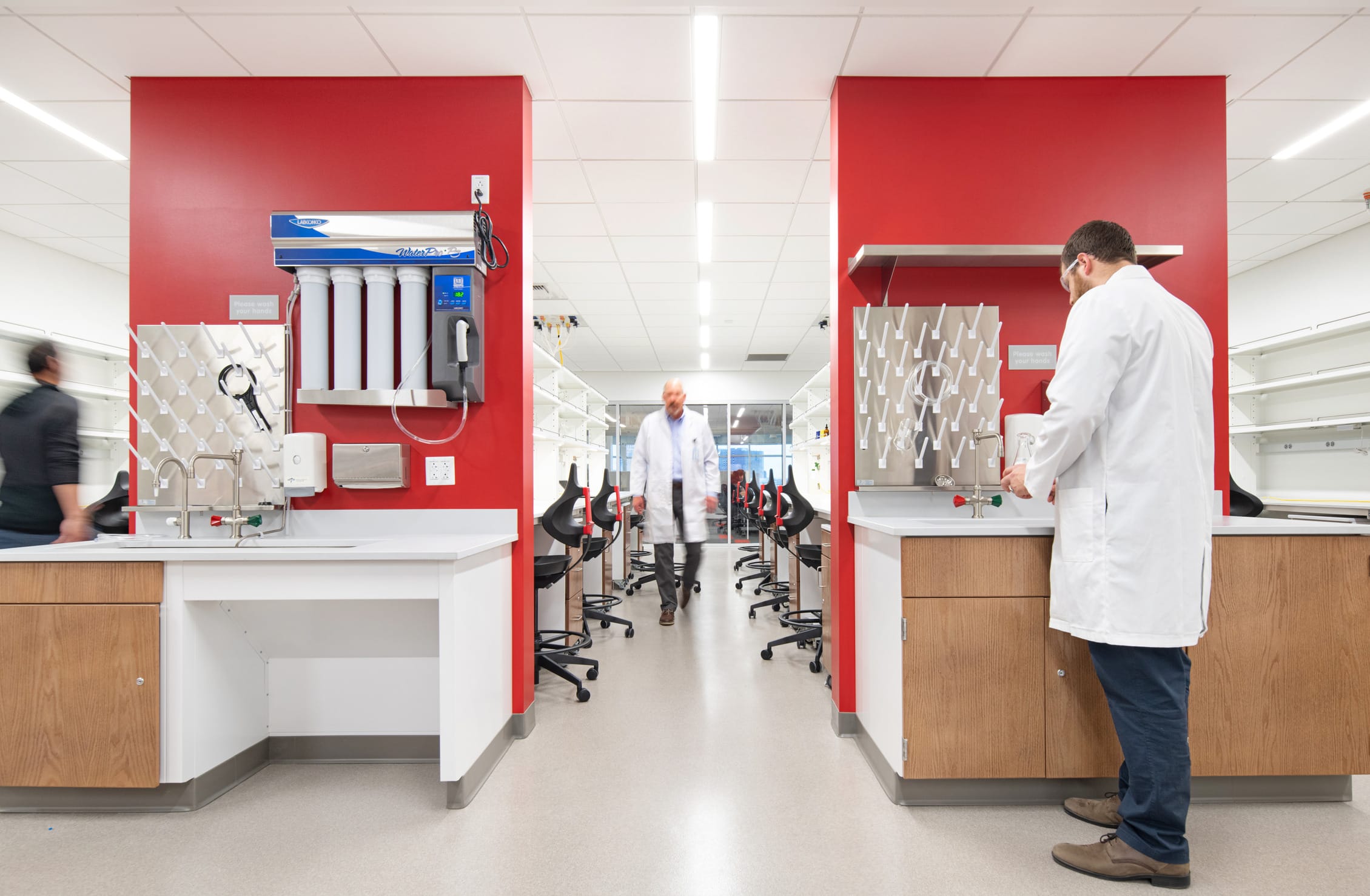 Laboratory workspace at Children's Mercy Research Institute featuring vibrant red accent walls that frame the entrance to an open lab area. On either side are workstations with white countertops and wooden cabinetry, equipped with sinks, water purification systems, and laboratory equipment. A researcher in a white lab coat works at one station while another lab-coated figure walks through the central corridor lined with black lab stools. The clean, modern design balances functional research requirements with distinctive architectural elements, showcasing the institute's state-of-the-art scientific facilities.