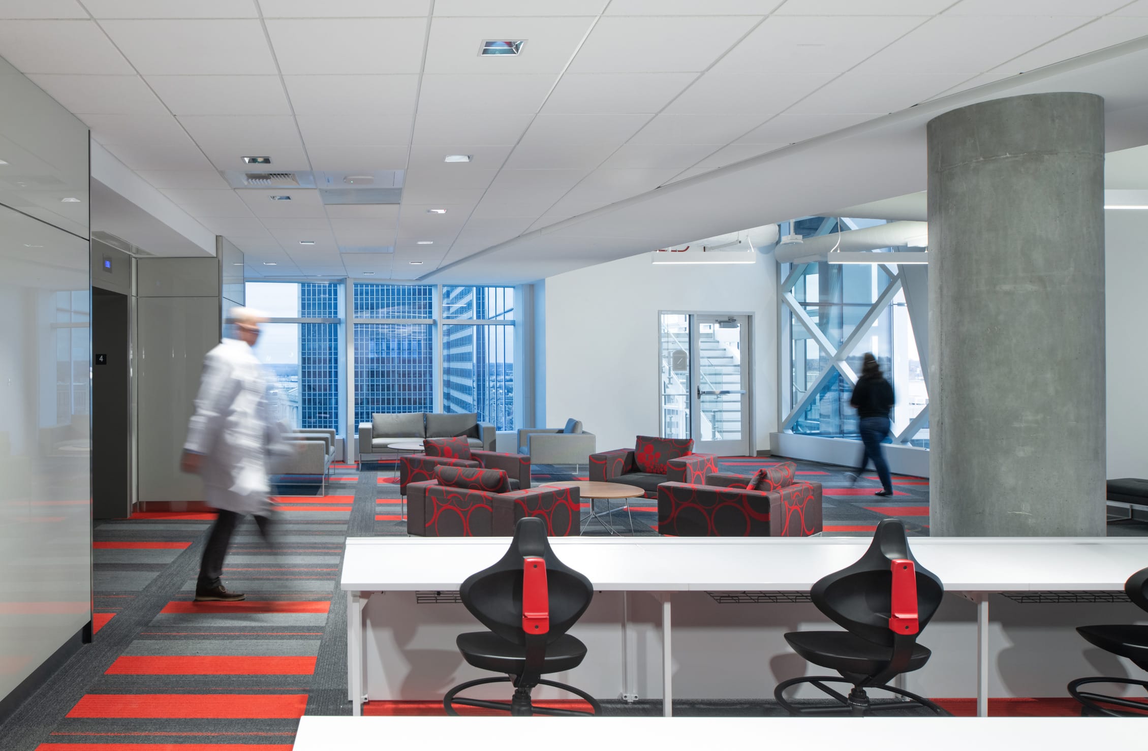 Collaborative workspace at the Children's Mercy Research Institute featuring a white lab table with black and red ergonomic stools on patterned gray and red carpet in the foreground. Beyond the table is a lounge area with red patterned accent chairs and grey couches near floor-to-ceiling windows offering panoramic views of Kansas City's skyline. Exposed concrete columns and elevator bays blend architectural elements with functional research spaces designed for both focused work and interaction.