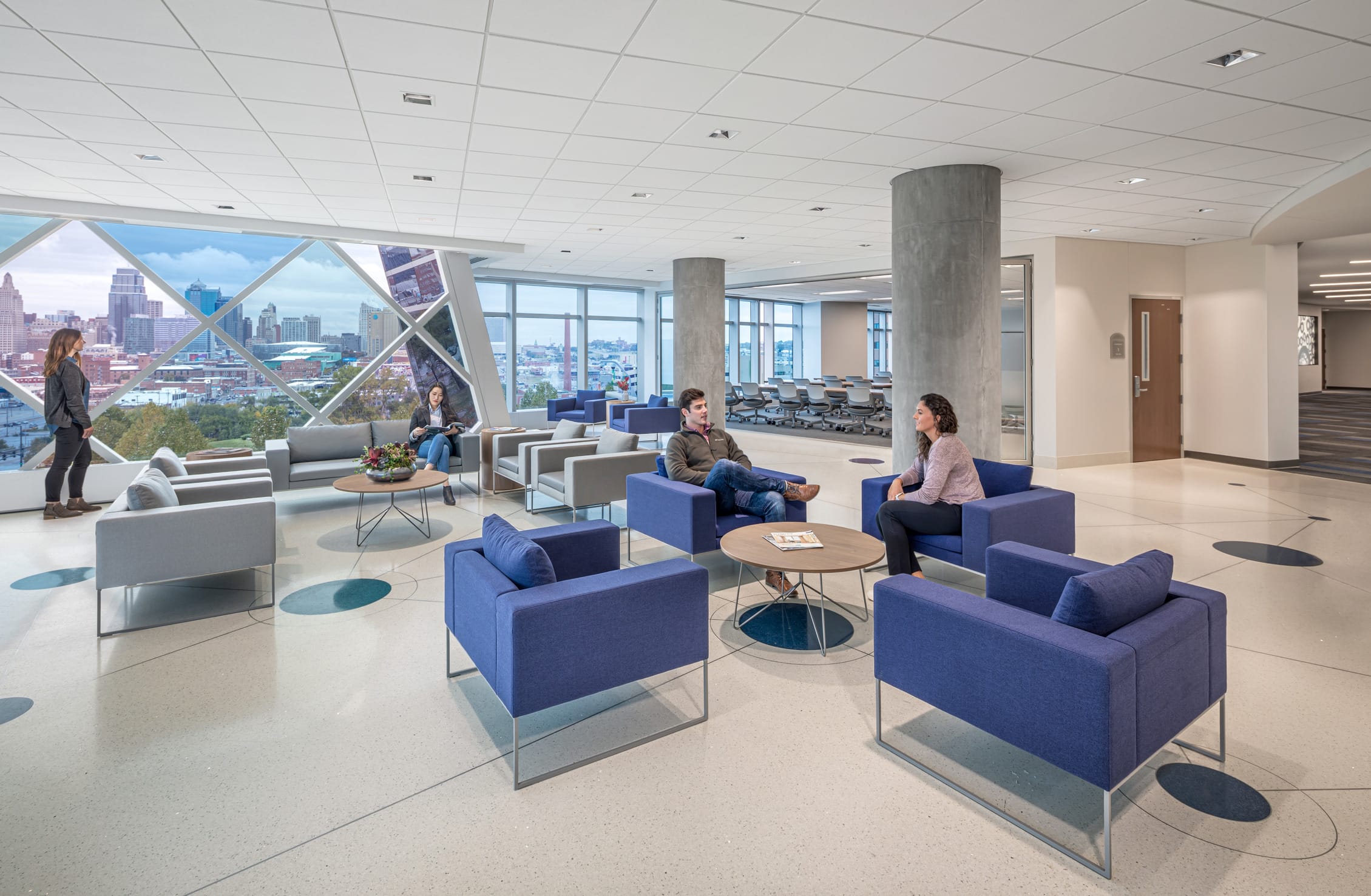 Bright communal lounge at the Children's Mercy Research Institute featuring floor-to-ceiling windows with distinctive diamond-pattern framing and transparent films to reflect different shades of blue, teal, and purple while also showcasing panoramic views of the Kansas City skyline. The spacious interior includes modern blue and gray seating arrangements with round wooden coffee tables, where researchers are engaged in conversation. Concrete columns punctuate the clean white space with subtle blue circular floor patterns. The thoughtfully designed environment balances professional functionality with comfortable areas for collaboration while maximizing natural light and city views.