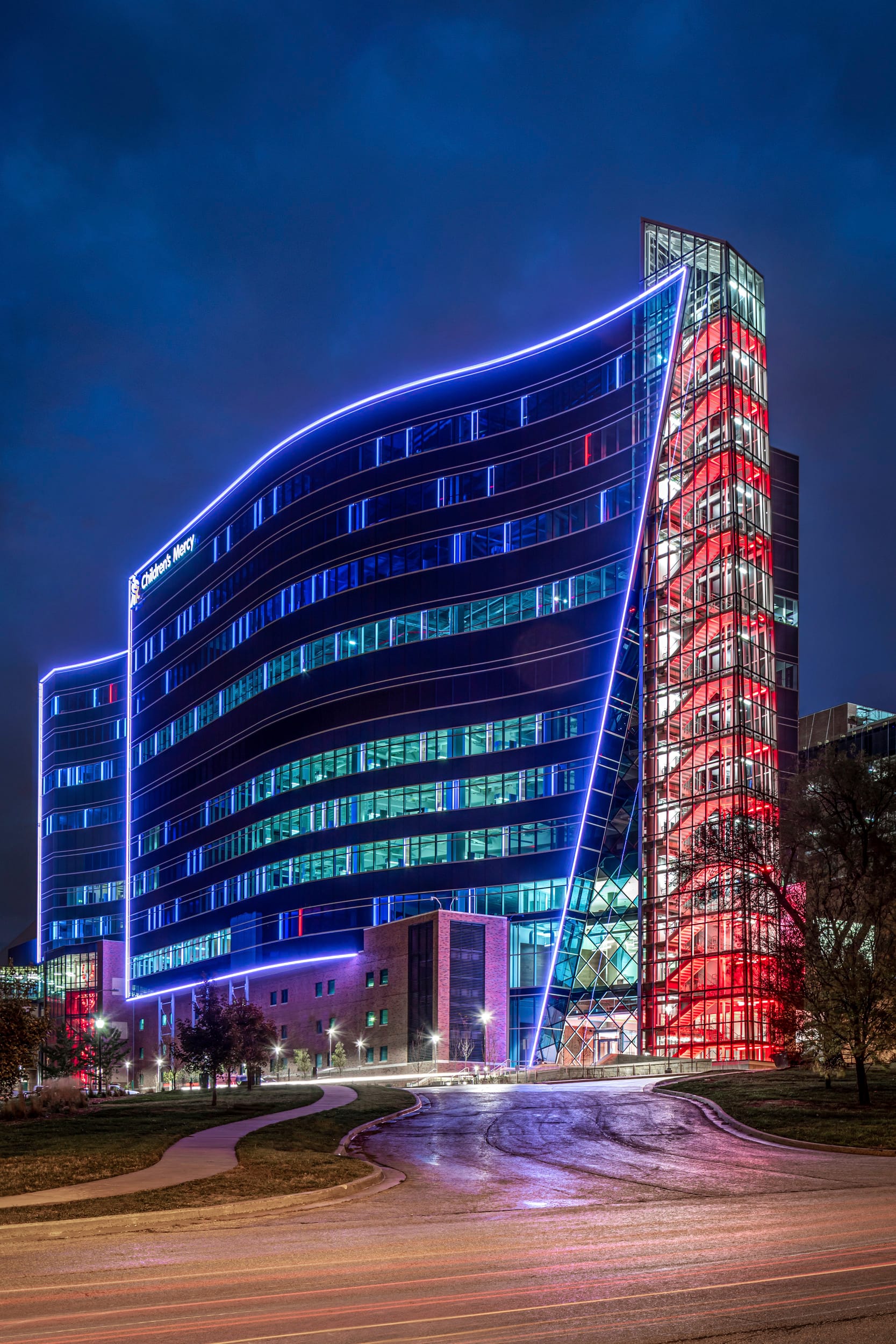 Night view of the Children's Mercy Research Institute, featuring a striking modern façade with curved blue LED lighting outlining the main glass structure. The building's distinctive design includes a prominent glass tower illuminated in vibrant red on the right side, creating a dramatic contrast against the deep blue night sky. The multi-story research facility showcases extensive glazing with interior lights visible through horizontal bands of windows, while curved walkways approach the entrance surrounded by landscaped grounds. Lighting patterns on the building are representative of DNA sequencing.