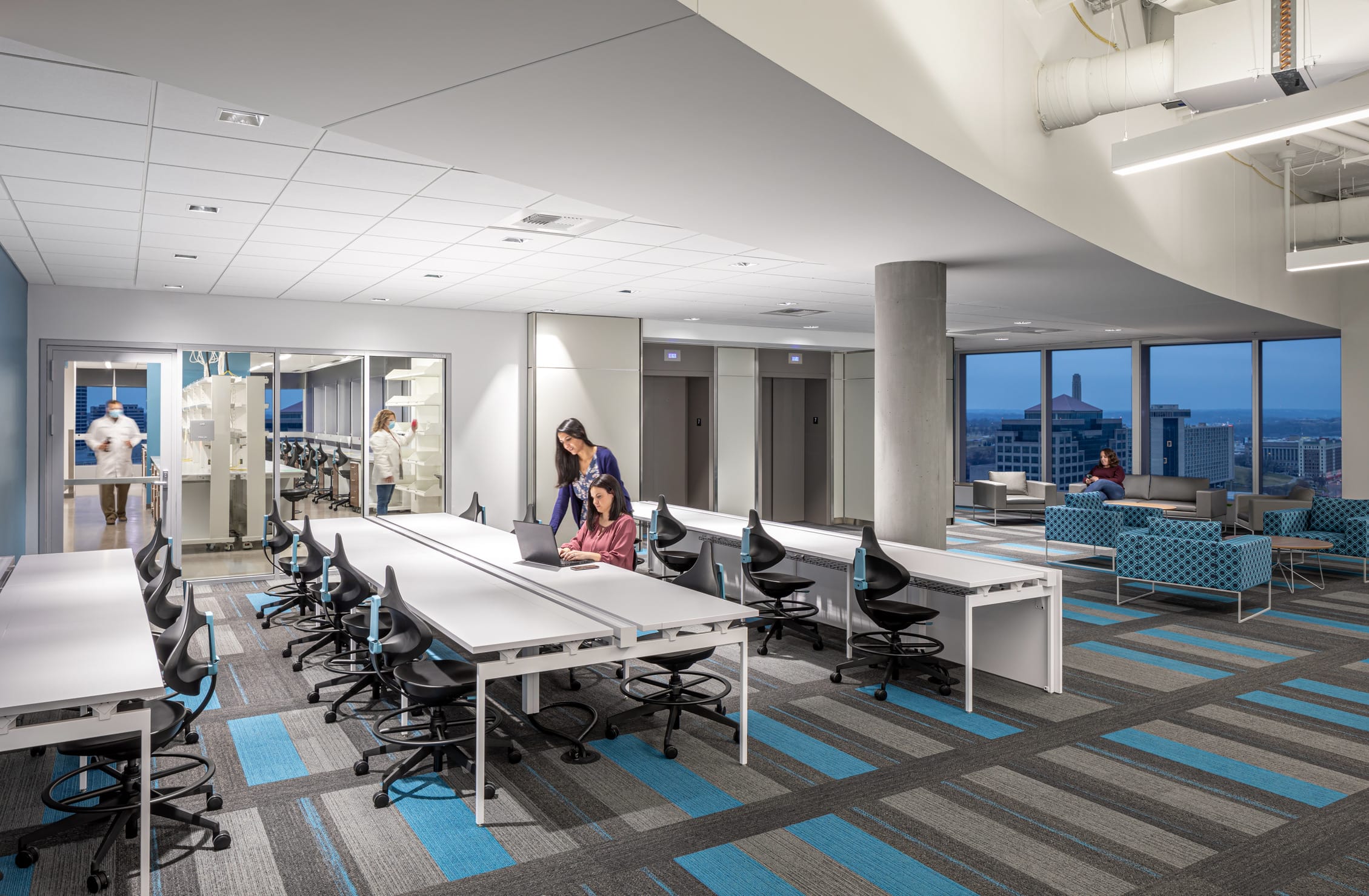 Collaborative workspace at the Children's Mercy Research Institute featuring white lab tables with black ergonomic stools arranged in rows on patterned gray and blue carpet. Two researchers work together at a laptop while laboratory spaces with glass walls are visible in the background where staff in white coats can be seen. The open floor plan transitions to a lounge area with blue seating near floor-to-ceiling windows offering panoramic views of Kansas City's skyline at dusk. Exposed concrete columns and elevator bays blend architectural elements with functional research spaces designed for both focused work and interaction.