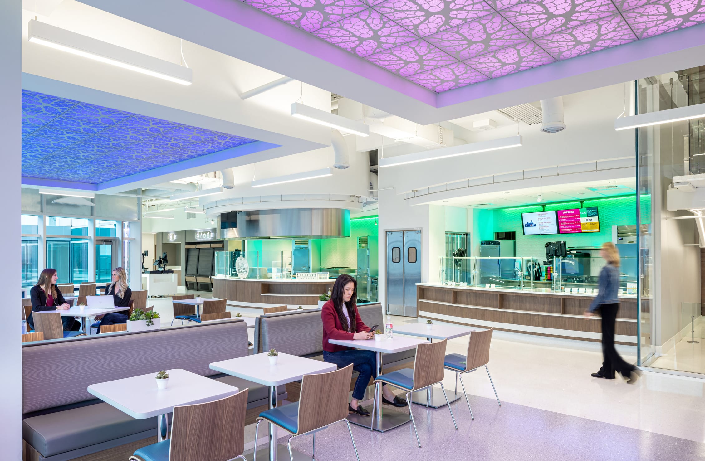 Modern cafeteria at the Children's Mercy Research Institute featuring illuminated ceiling panels with cellular patterns in purple and blue. The bright, open dining area includes white tables with wooden chairs and built-in banquette seating where several people are seated. One woman works on a laptop while two others converse at a nearby table. The contemporary serving area is visible with green ambient lighting, digital menu displays, and stainless steel serving stations, creating a welcoming space that blends scientific design elements with functional dining amenities.