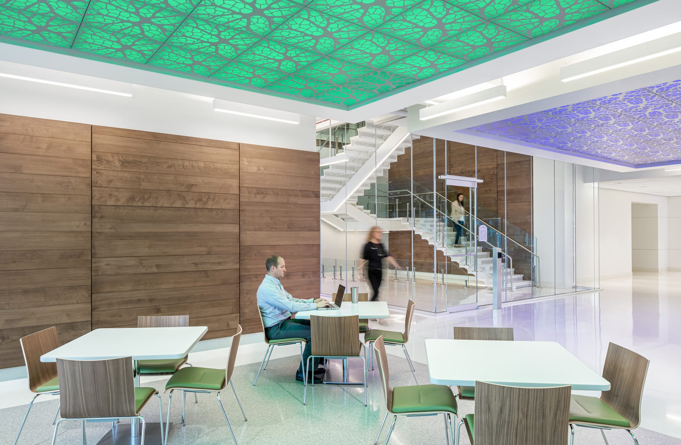 Interior collaborative space at the Children's Mercy Research Institute featuring warm wood wall panels contrasted with modern white stairs and glass railings. A researcher works on a laptop at one of several white tables with wooden and green-cushioned chairs. The space is distinguished by decorative illuminated ceiling panels in green and blue with cellular patterns. Two people are in motion on the central staircase, highlighting the dynamic nature of this bright, open workspace designed to facilitate interaction.