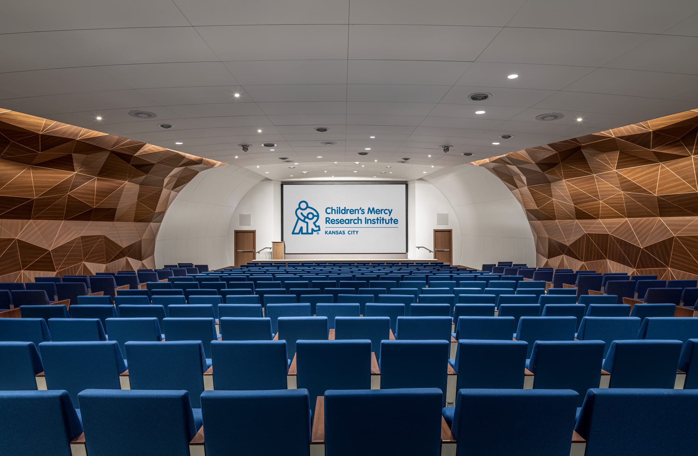 A modern auditorium at the Children's Mercy Research Institute in Kansas City, designed by BSA, featuring rows of vibrant blue seating facing a large presentation screen. The space is defined by its sculptural wooden acoustic panels with geometric patterns and curved white walls, creating a visually striking and acoustically refined environment for lectures and events.