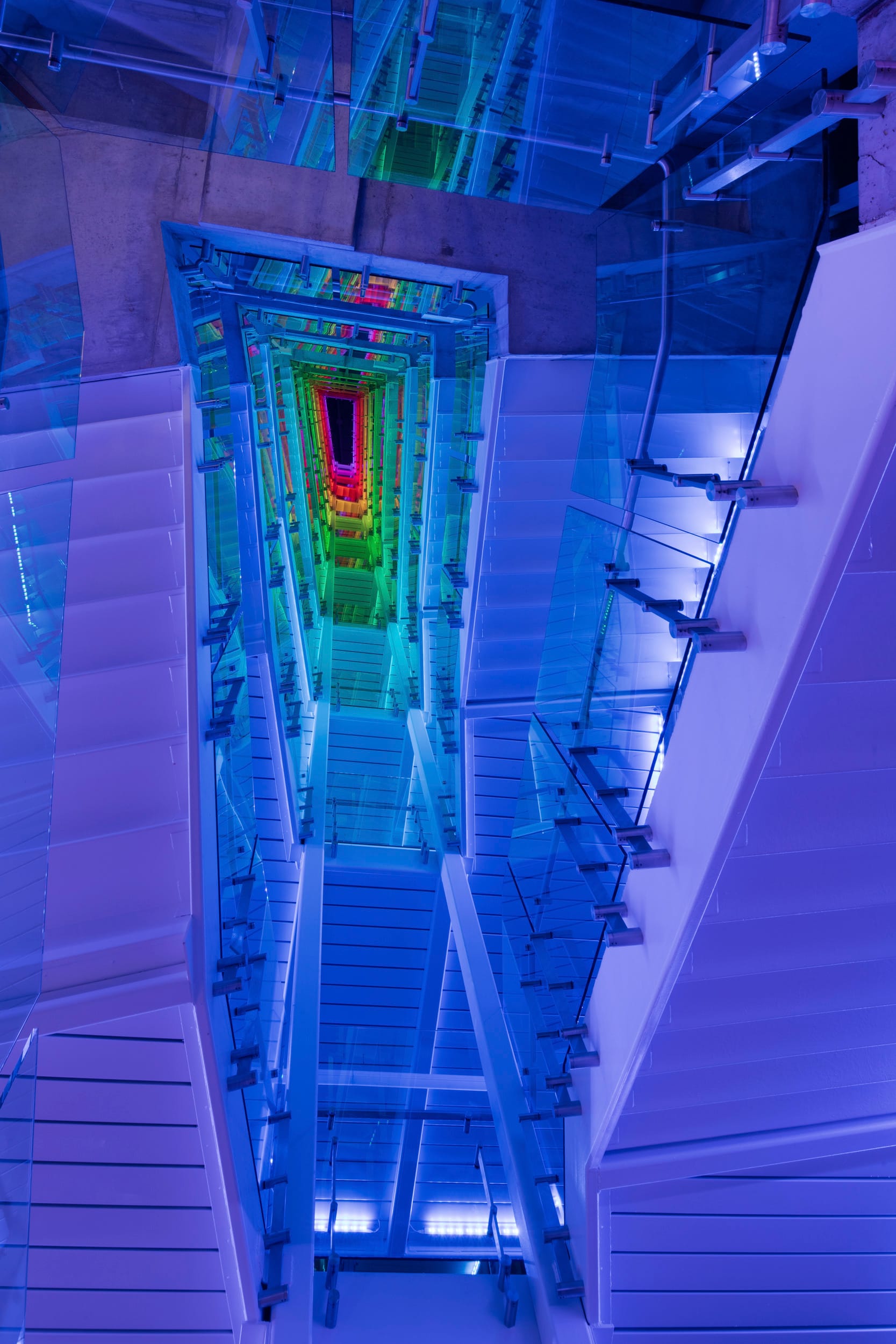 Looking up through a dramatic multi-story stairwell at the Children's Mercy Research Institute, designed by BSA, where vivid LED lighting shifts from deep purple to vibrant rainbow hues as it ascends. Glass railings and sleek white walls enhance the futuristic and immersive visual effect of the space.