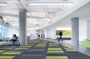 A bright, modern interior of the Children's Mercy Research Institute, designed by BSA, featuring bold green and gray patterned carpeting and matching green patterned accent chairs and wall decor. The open layout includes a casual seating area, workstations, and cubicle-like offices in the back, creating a welcoming and vibrant research environment. Large concrete pillars frame the image.