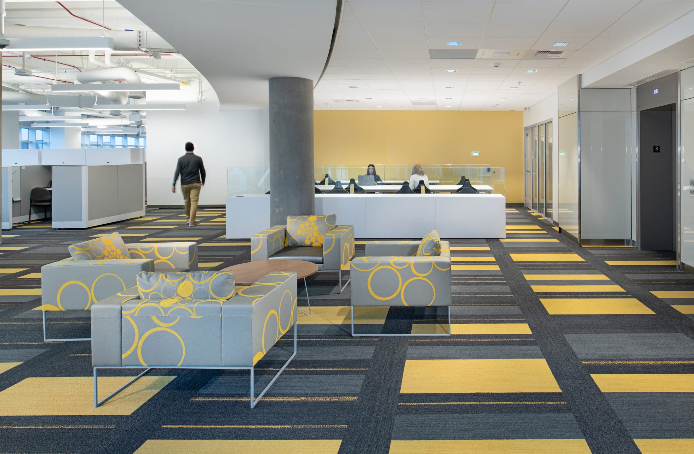 A bright, modern interior of the Children's Mercy Research Institute, designed by BSA, featuring bold yellow and gray patterned carpeting and matching lounge chairs. The open layout includes a casual seating area, workstations, and a sleek white reception desk set against a yellow accent wall, creating a welcoming and vibrant research environment.