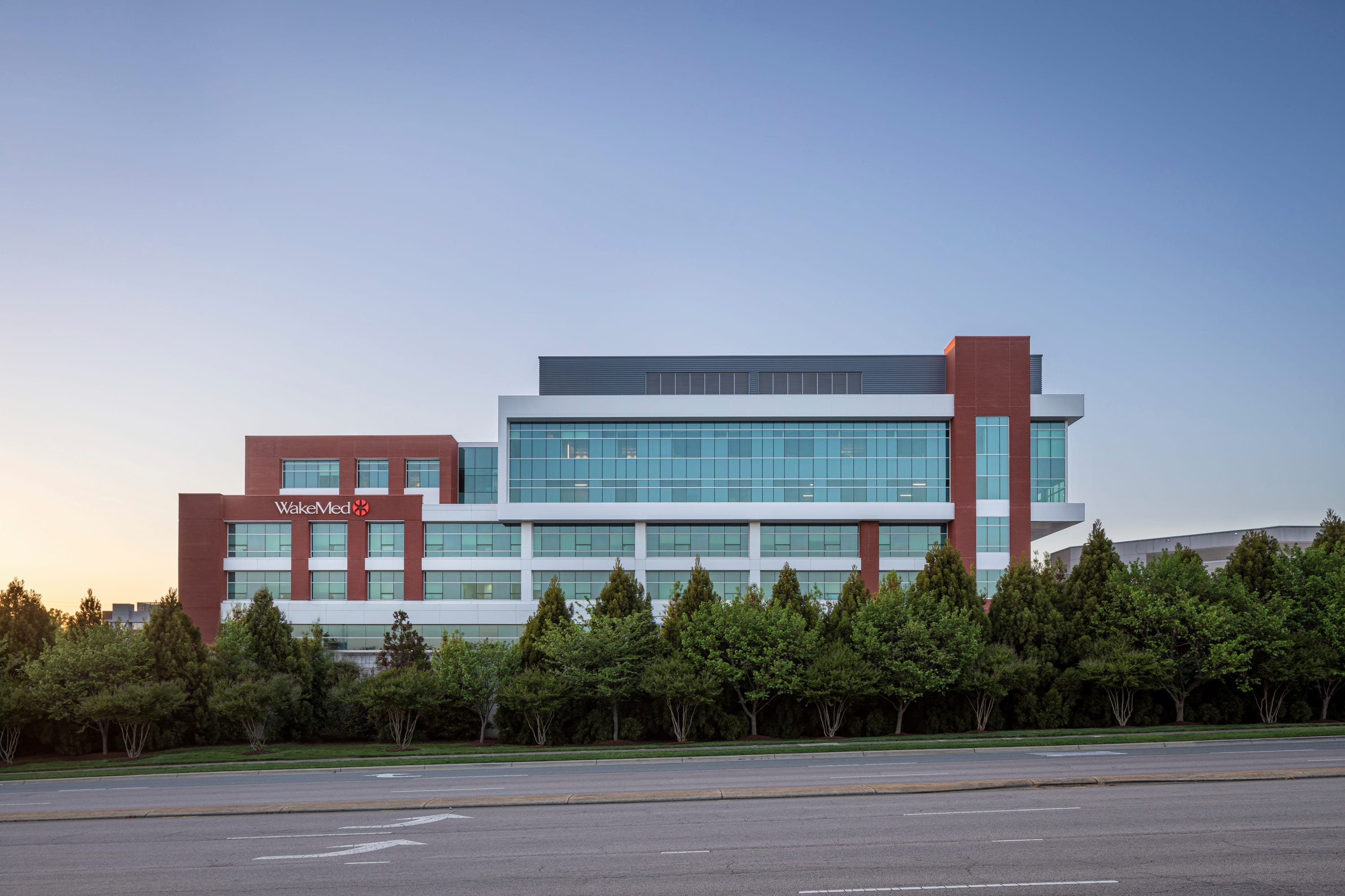 View of the WakeMed Cary Hospital, a modern medical facility building with red brick and light metal exterior with large glass windows from the highway. The building has a 'WakeMed' logo on the brick and is against a clear blue sky at dawn or dusk.