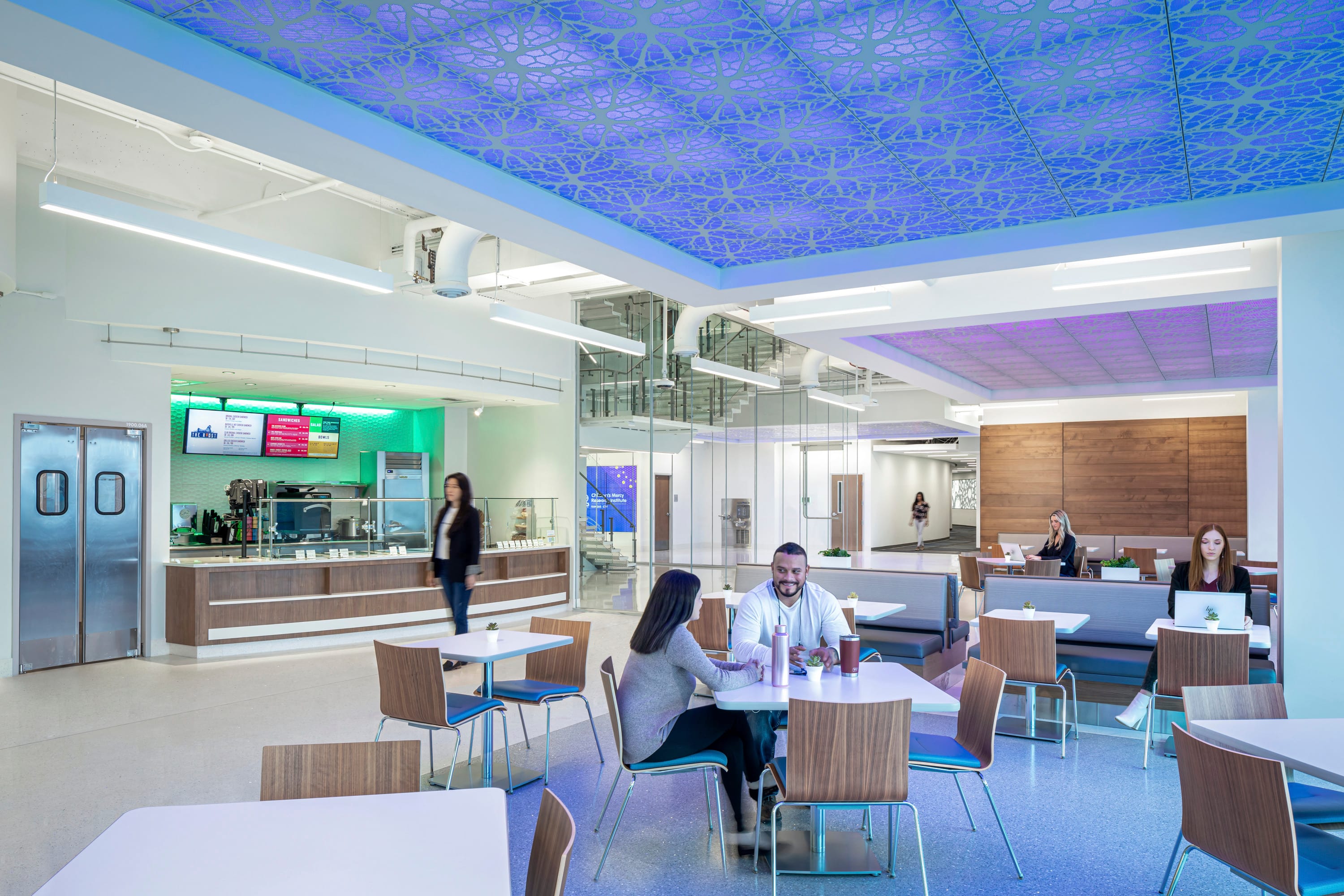 Modern cafeteria at the Children's Mercy Research Institute featuring illuminated ceiling panels with cellular patterns in purple and blue. The bright, open dining area includes white tables with wooden chairs and built-in banquette seating where several people are seated. The contemporary serving area is visible with green ambient lighting, digital menu displays, and stainless steel serving stations, creating a welcoming space that blends scientific design elements with functional dining amenities.
