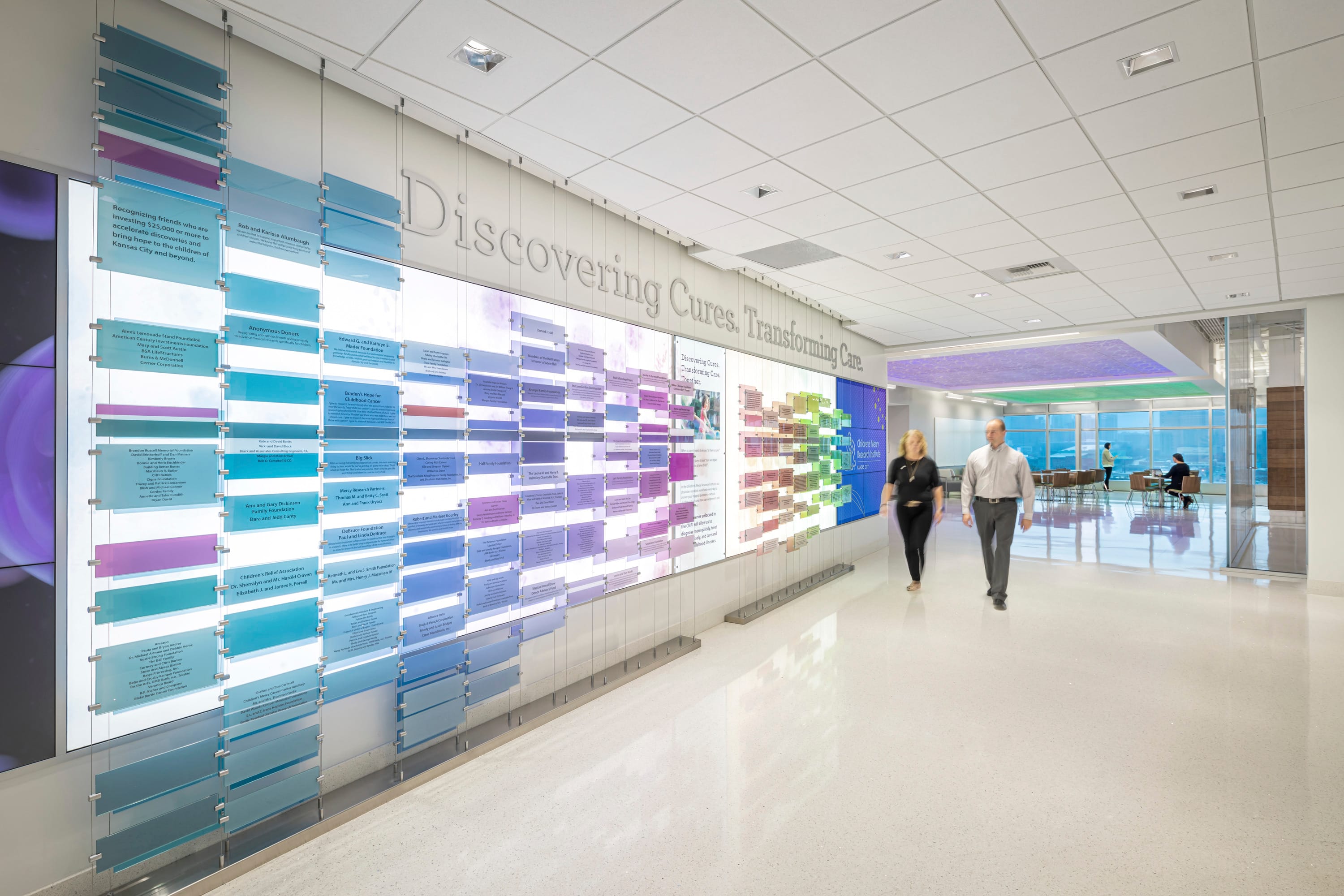 Bright corridor in the Children's Mercy Research Institute featuring an illuminated donor recognition wall with the motto 'Discovering Cures, Transforming Care' displayed prominently. The wall consists of numerous blue and purple acrylic panels arranged in horizontal rows, containing names and information about supporters. Two staff members walk through the polished hallway, while a meeting space with large windows providing natural light is visible in the background. The modern, airy design embodies the institute's commitment to transparency in both architecture and mission.