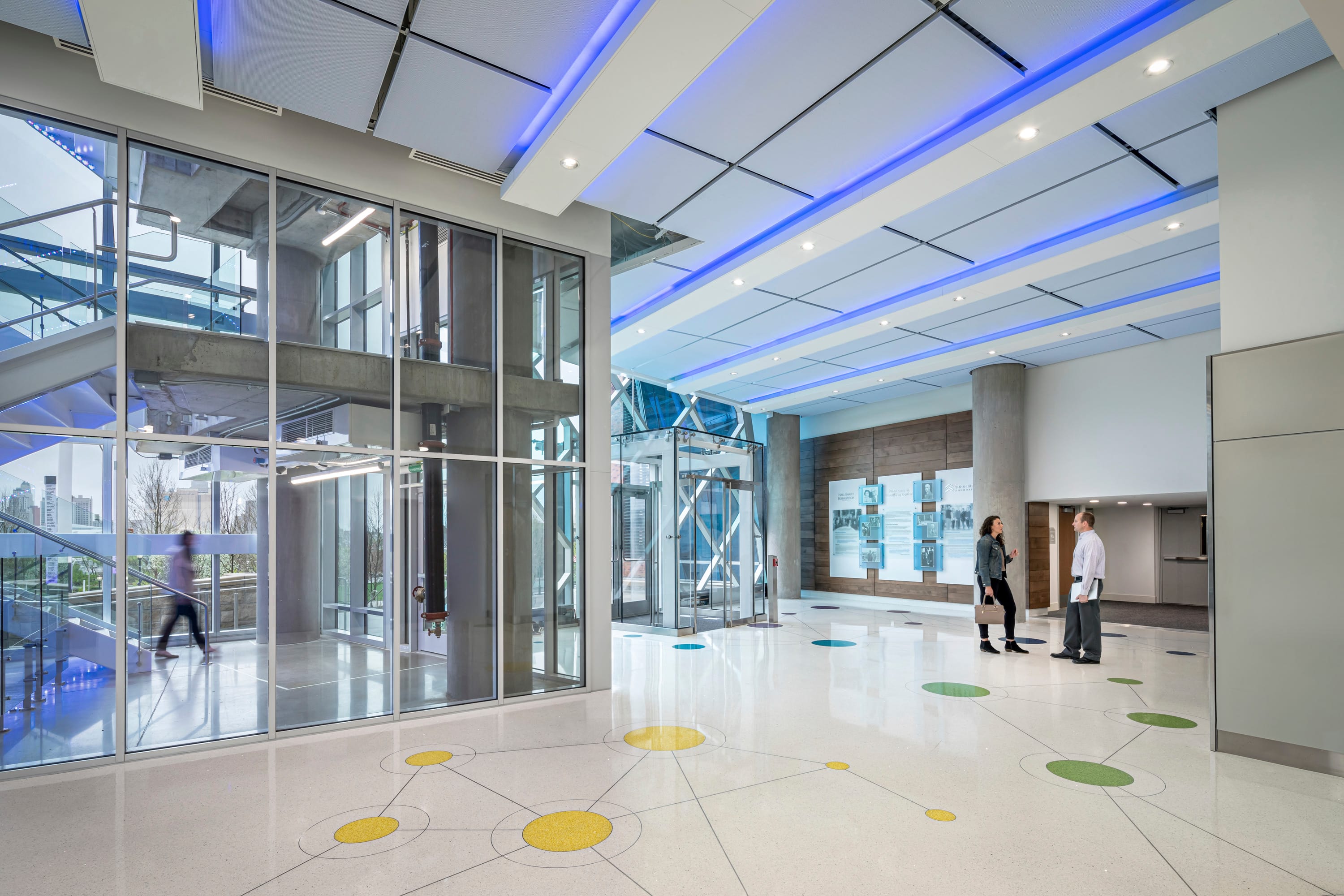 This image shows the modern interior lobby of what appears to be the Children's Mercy Research Institute. The space features a sleek design with a white polished floor decorated with colorful circular patterns in yellow and green. The ceiling has blue LED accent lighting running along white panels. Large glass walls and windows create an open, airy atmosphere, revealing structural concrete elements. In the foreground, two people are conversing - a woman in dark clothing with a bag and a man in a white shirt. The lobby includes information displays on a wood-paneled wall, an elevator or glass enclosure, and hints of an urban skyline visible through windows. The overall aesthetic combines contemporary architecture with bright, welcoming elements appropriate for a research facility.