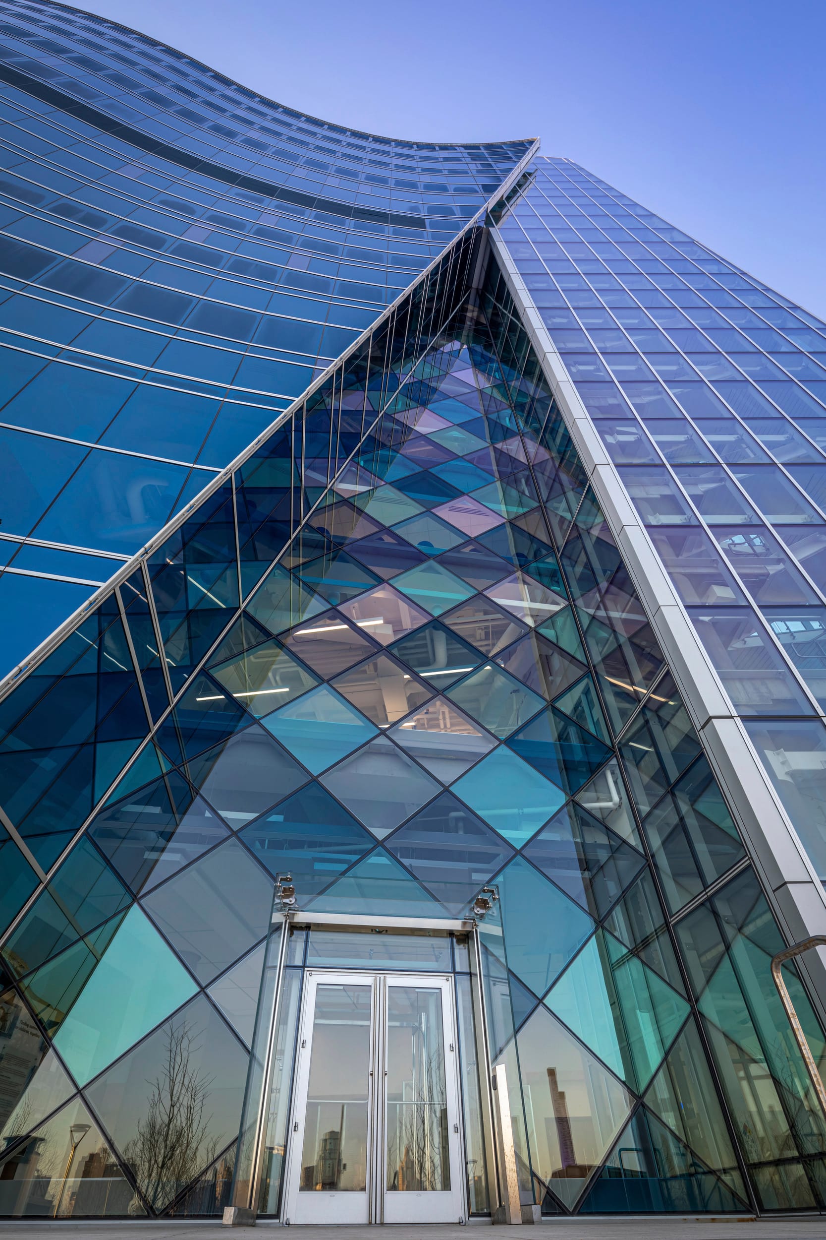 This image shows an upward-looking view of a modern architectural entrance to the Children's Mercy Research Institute. The entrance features a striking glass atrium with a triangular or diamond-patterned façade that incorporates various shades of blue, purple, and teal glass panels. Above the entrance, the building rises in a curved form with extensive blue-tinted glass curtain walls that reflect the sky. The glass entrance doors are set within this geometric glass feature, creating a dramatic and visually interesting entryway. The perspective is from ground level looking up, emphasizing the height and modern design of the building against a clear blue sky.