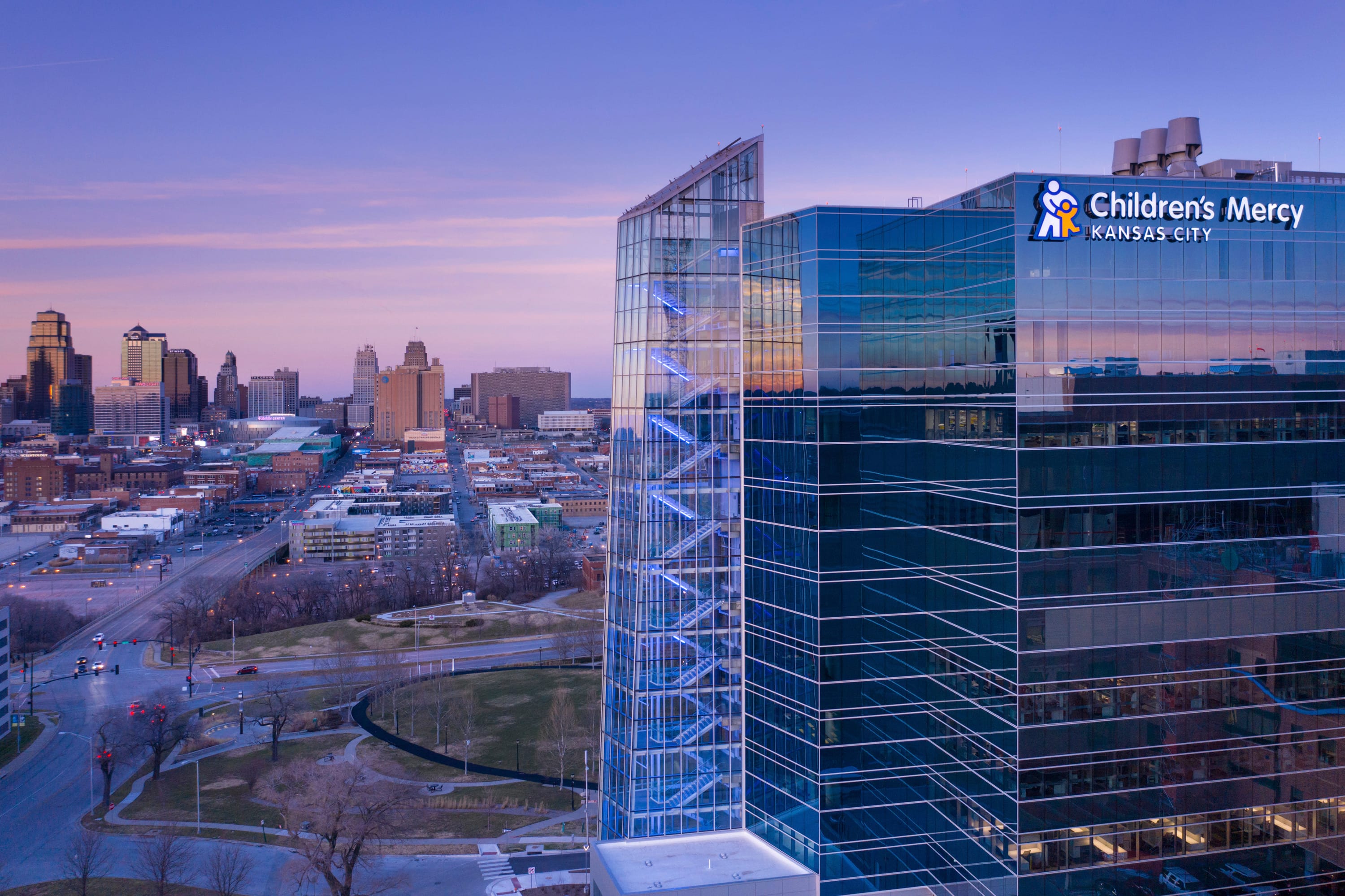 This image shows the exterior of the Children's Mercy Kansas City hospital building during what appears to be dusk or early evening. The modern, entirely glass building features the Children's Mercy logo (with a figure of an adult holding a child) prominently displayed on the upper right corner. The structure has a distinctive blue-tinted glass façade with illuminated blue accents visible along an all-glass stairwell tower on the left side of the building. In the background, you can see the Kansas City skyline with various high-rise buildings. The foreground shows roads, some bare trees, and open spaces surrounding the hospital, suggesting it's during winter or early spring. The purple-blue sky creates a dramatic backdrop for both the hospital and the city skyline.