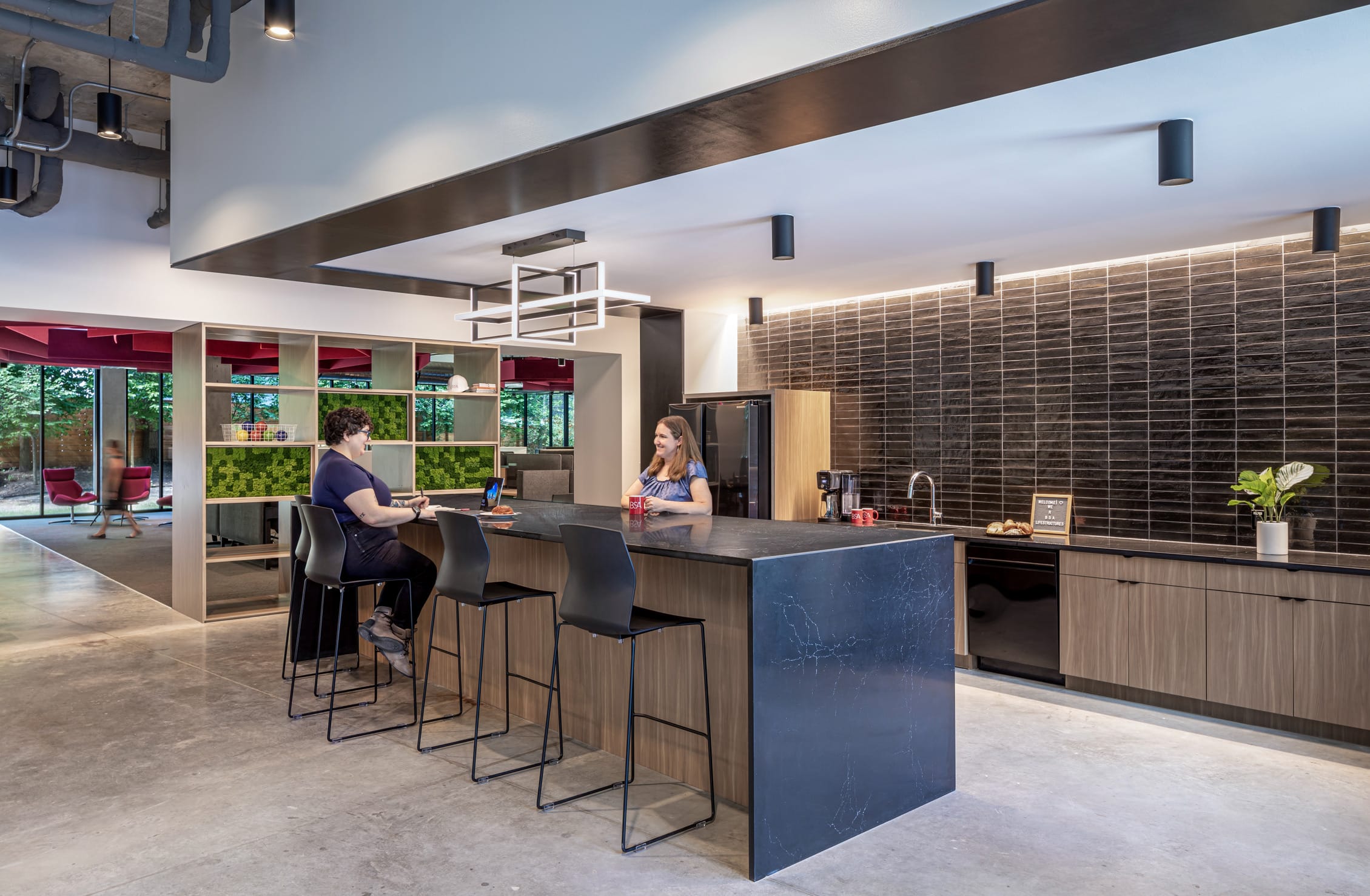 This image shows a modern office break area with a sleek kitchen design. Two people are conversing at a stylish kitchen island with dark stone countertops and wood paneling, complemented by minimalist black bar stools. The kitchen features dark tile backsplash, built-in appliances, and contemporary fixtures including a geometric pendant light. The space opens to a collaborative area with bookshelf dividers incorporating green plant panels, red accent seating, and large windows providing natural light. The design showcases an industrial-modern aesthetic with exposed ceiling elements, concrete flooring, and a thoughtful balance of materials and lighting that creates a welcoming, functional workspace environment.
