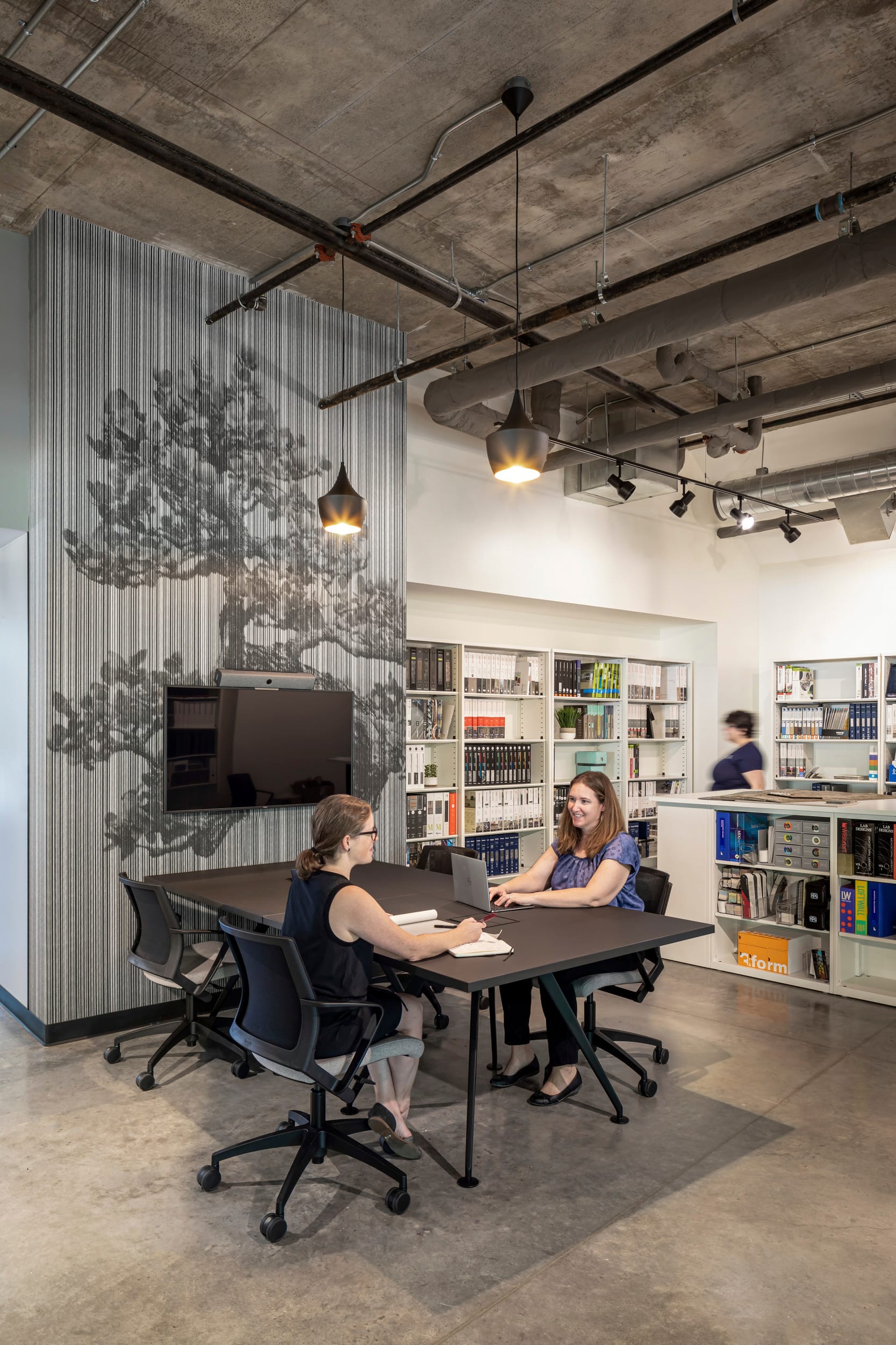 This image showcases a collaborative meeting space in BSA's office where two professionals engage in discussion at a dark wood conference table with ergonomic office chairs. The design features an artistic wall treatment with vertical striping and a subtle tree silhouette, creating visual interest while maintaining the sophisticated atmosphere. Pendant lighting with metallic finishes hangs from an exposed concrete ceiling that reveals the building's structural and mechanical systems. In the background, extensive white bookshelves house the firm's reference materials and resources, while polished concrete flooring completes the modern, industrial aesthetic. This thoughtfully designed meeting area exemplifies BSA's commitment to creating spaces that balance functionality with distinctive design elements that inspire creative collaboration.