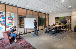 This image showcases BSA's versatile conference area designed to support multiple work modes simultaneously. In the foreground, a person sits in a coral accent chair on a colorful patterned rug, observing a colleague who is sketching ideas on a mobile whiteboard. The background reveals the main conference room where three professionals collaborate around a wooden conference table with black ergonomic chairs on a defined area rug. The space features BSA's signature design elements: wood and glass partitions with black frames, concrete flooring, integrated technology including a wall-mounted display, and built-in storage with green moss panel accents. A vibrant, colorful mural is visible through the glass partition, adding artistic interest to the professional environment. This thoughtfully designed space demonstrates BSA's expertise in creating flexible, collaborative work environments that support various meeting styles and creative processes.