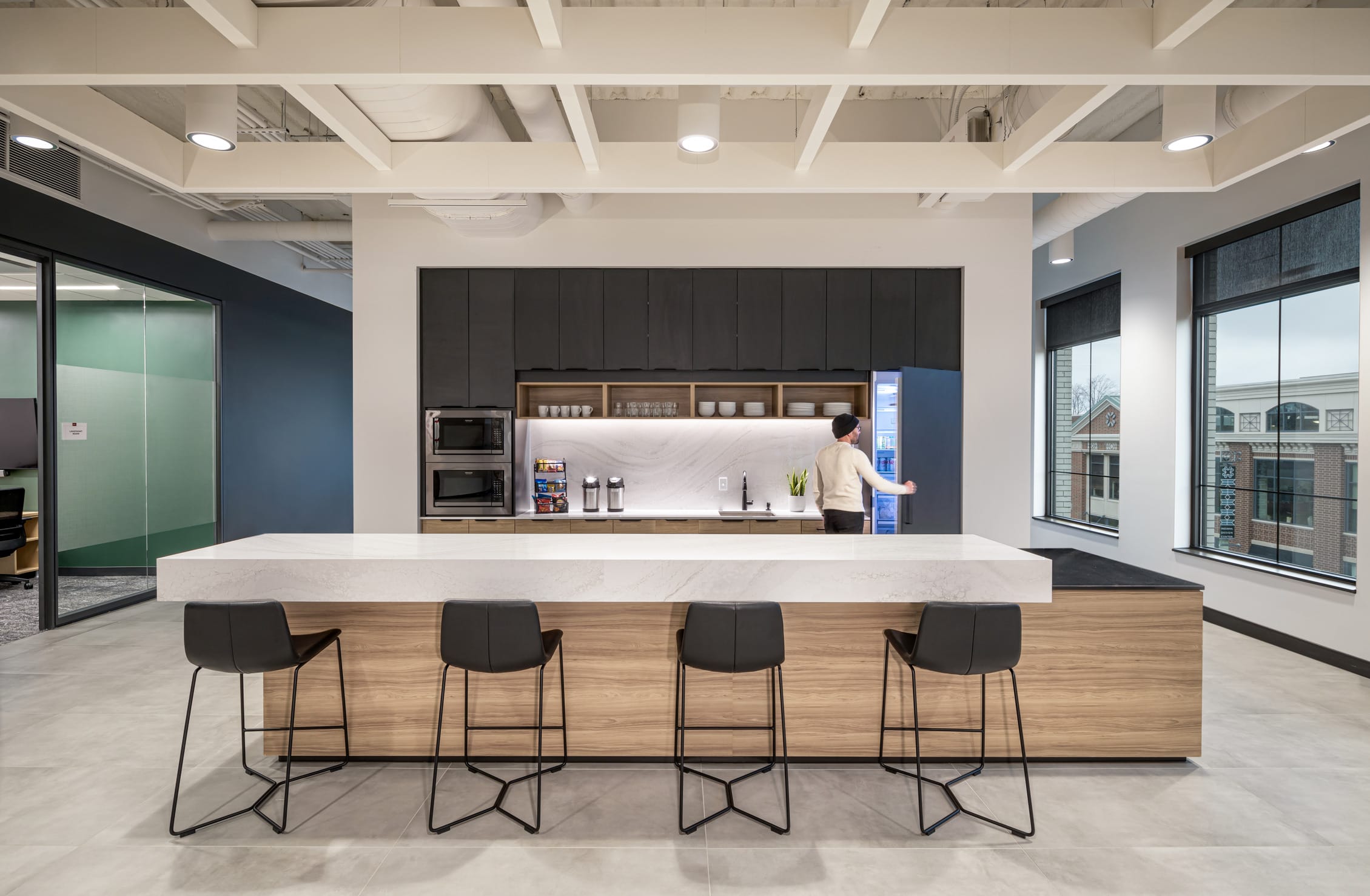 This image features BSA's modern office kitchen and break area, designed with both functionality and style. The space centers around a large island with a substantial white quartz countertop supported by warm wood cabinetry, with four contemporary black bar stools arranged along the front. Behind the island is a well-equipped kitchenette featuring dark gray upper cabinets, open shelving displaying dishware, integrated appliances, and a backlit white marble backsplash. A person can be seen accessing the refrigerator. The space benefits from natural light through large windows revealing neighboring buildings, and features an exposed ceiling with white structural elements, recessed lighting, and light-colored flooring. Adjacent glass-walled meeting spaces with accent walls are visible, highlighting BSA's thoughtful approach to creating collaborative work environments.
