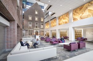 The stunning main lobby of Riley Hospital for Children's Maternity Tower showcases a dramatic multi-story atrium that seamlessly blends historic brick architecture with contemporary design elements. The space features a mix of white modern seating and plum-colored armchairs arranged throughout the expansive area, while dramatic golden wood grain panels create visual impact on the upper level walls. Natural light floods in through skylights above, illuminating the preserved historic brick façade that forms the centerpiece of this welcoming family-centered environment, with visitors and staff moving comfortably through the thoughtfully designed space.