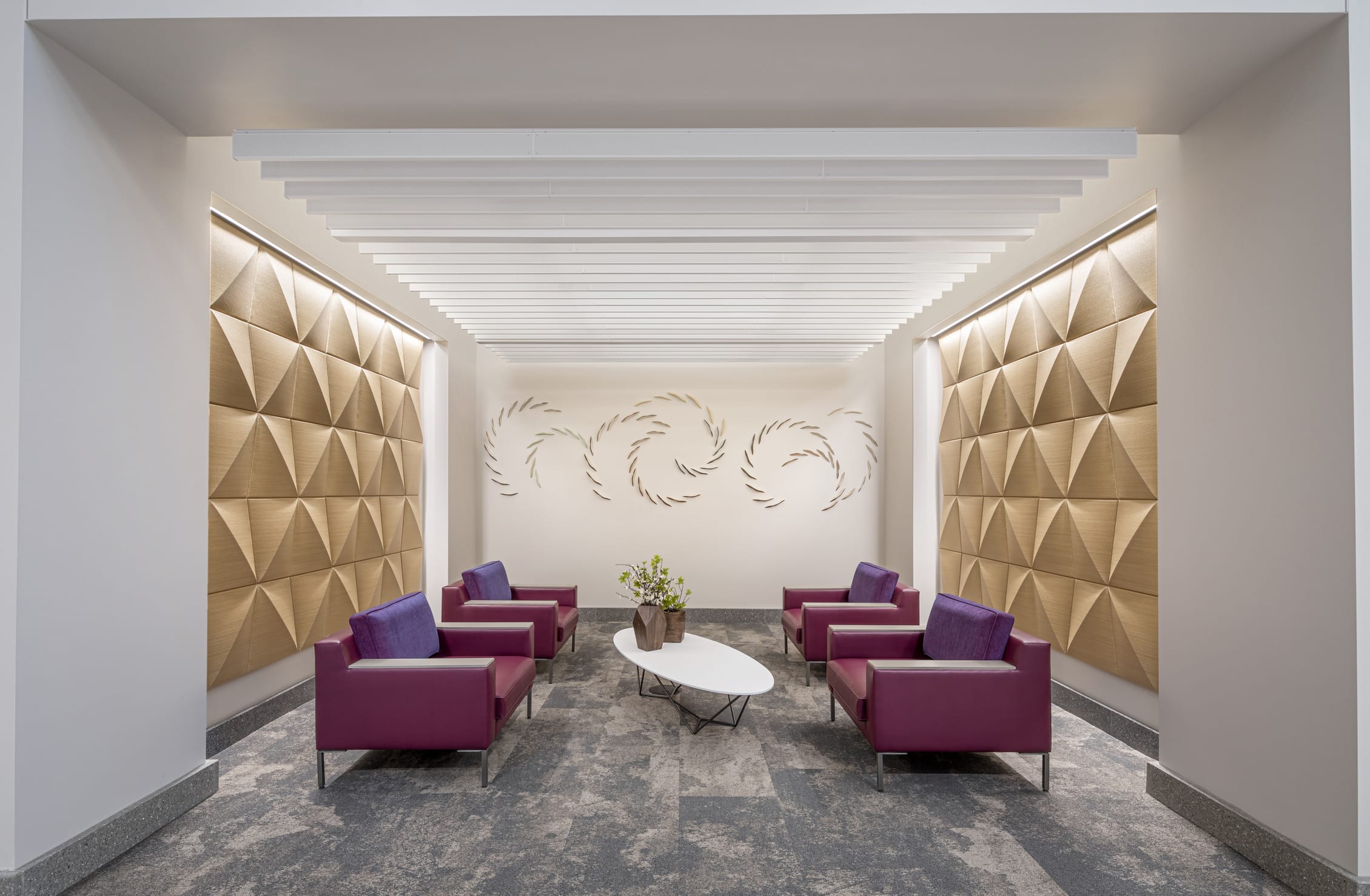 An elegant waiting area at Riley Hospital for Children's Maternity Tower features four plum-colored armchairs with purple accent pillows arranged around a white oval coffee table in an intimate seating alcove. The sophisticated space is enhanced by dramatic geometric wood paneling on both side walls, a striking linear ceiling design with integrated lighting, and decorative circular wall art, all set on polished stone flooring that creates a calming and upscale environment for expectant families.