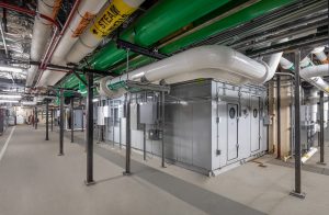 This image shows the mechanical equipment room at Riley Hospital for Children at IU Health Maternity Tower, featuring large industrial HVAC units and building systems infrastructure. The space contains multiple gray metal air handling units with circular inspection ports, surrounded by an extensive network of ductwork, piping, and conduits running along the ceiling. Green-colored pipes, white insulated ducts, and various colored utility lines create a complex mechanical systems layout typical of a modern hospital's building services area. The room has concrete flooring and support columns, with clear pathways for maintenance access around the equipment. This behind-the-scenes mechanical space is essential for providing climate control, ventilation, and other building systems that support the hospital's patient care areas.