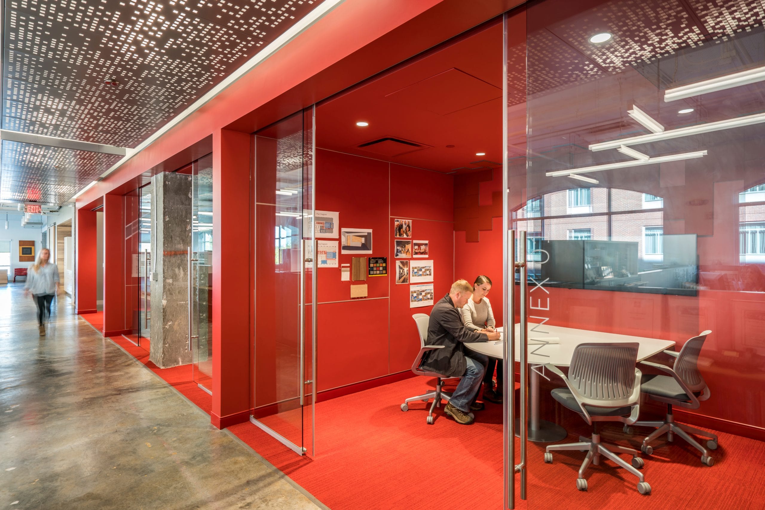 This image showcases BSA's innovative approach to office meeting spaces with a row of glass-enclosed huddle rooms featuring vibrant red interiors that create bold visual impact against the polished concrete corridor floor. In the foreground meeting room labeled
