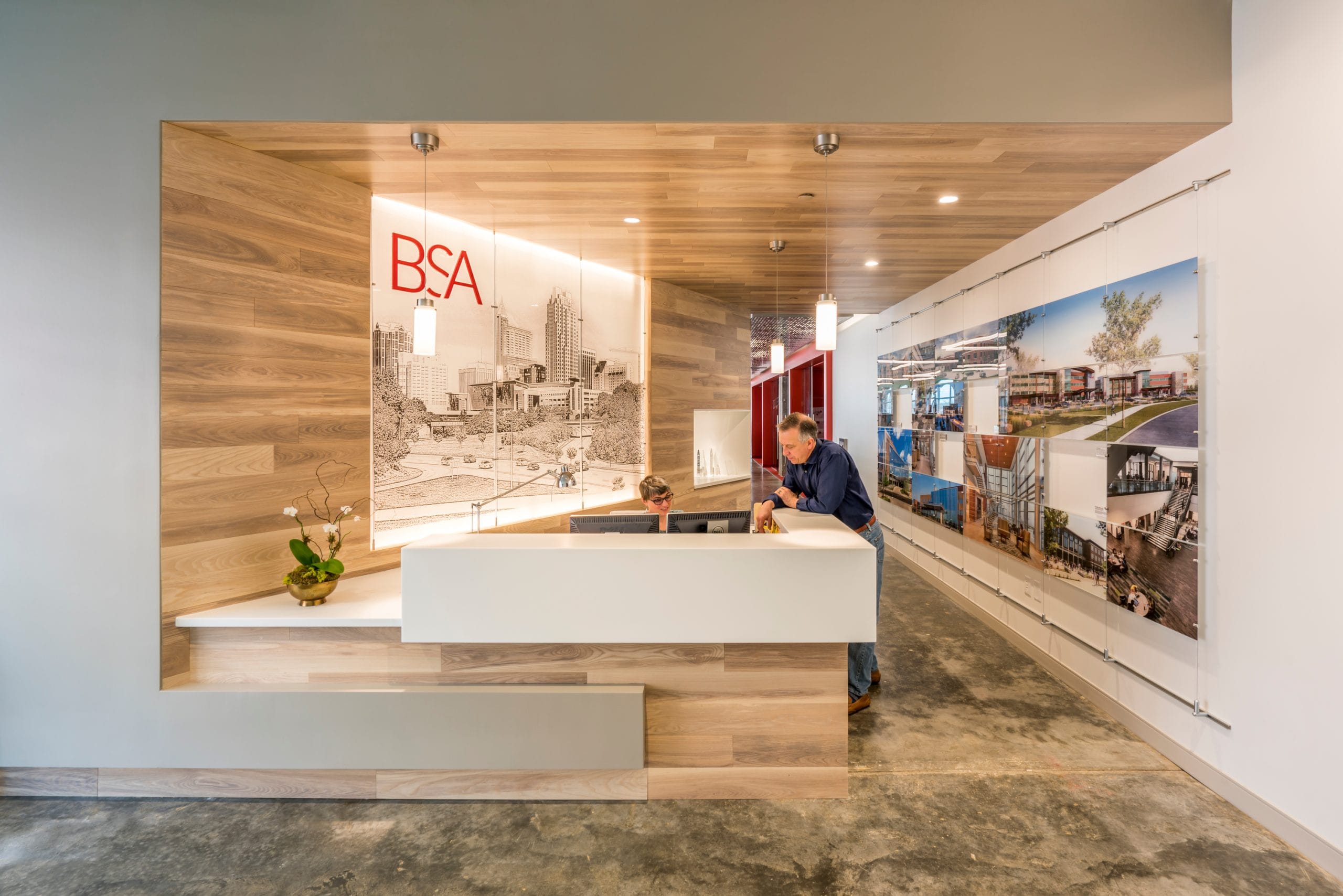 This image shows the elegant reception area of BSA, an architecture, engineering, and interior design firm. The modern space features a stylish white reception desk positioned within a warm wooden alcove with pendant lighting. Behind the desk is a backlit wall displaying