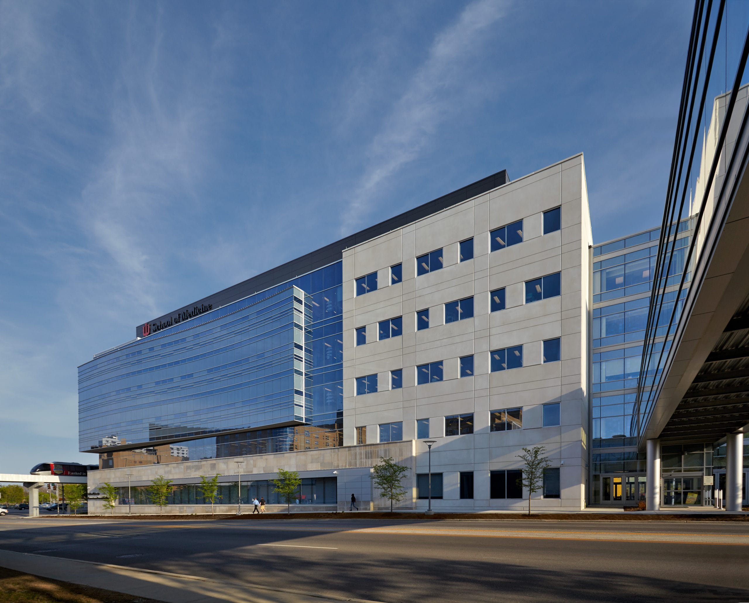 This image shows a modern building with a striking architectural design. The structure features a combination of materials, with a rectangular section made of light gray paneling or concrete on the right side, and a curved glass façade on the left that dramatically reflects the blue sky. The curved glass section creates a dynamic contrast against the more traditional rectangular portion of the building. There is a glass enclosed pedestrian bridge or urban skyway connecting the building to a structure that cannot be seen in this image.