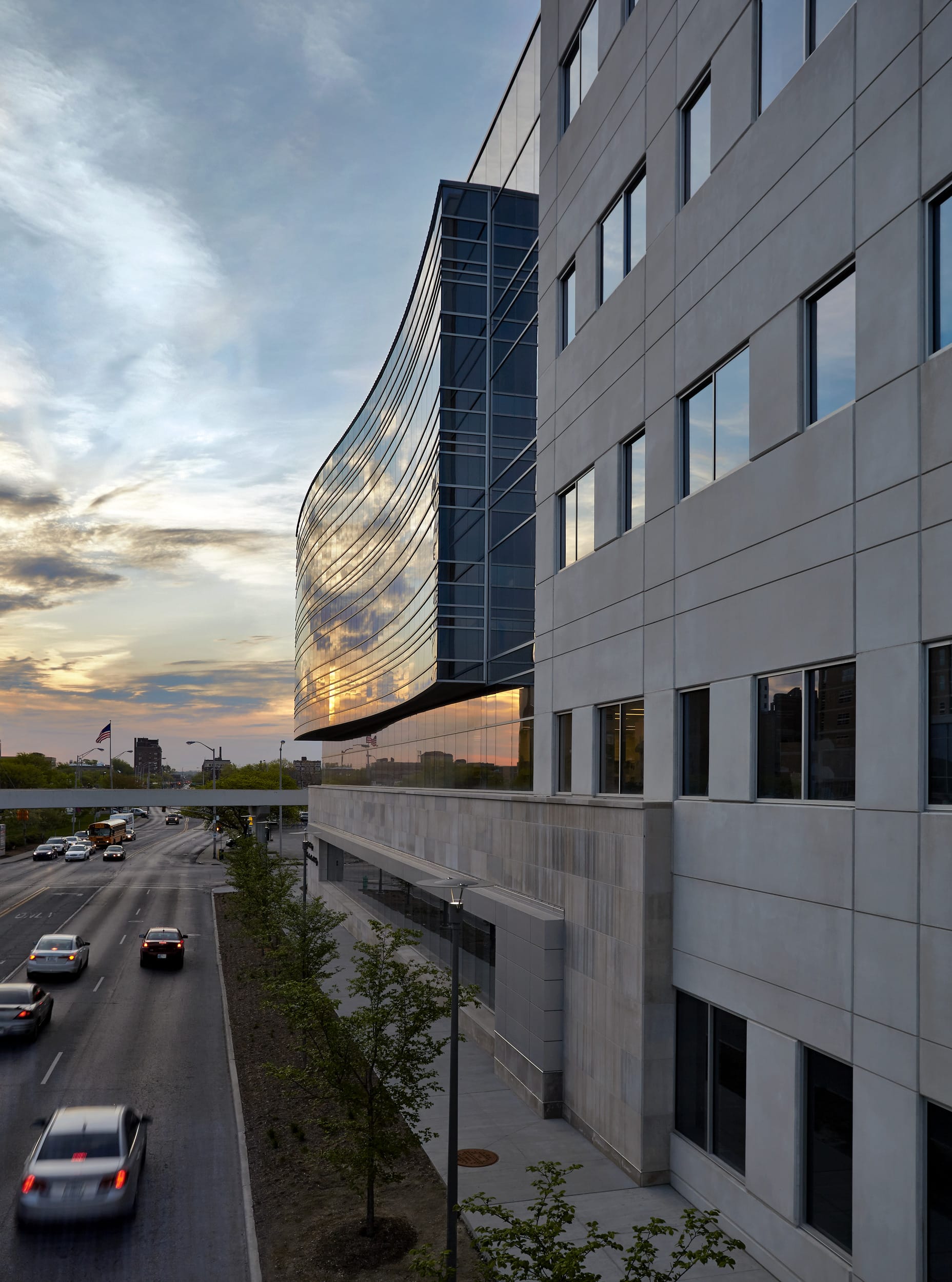 This image shows a modern building with a striking architectural design. The structure features a combination of materials, with a rectangular section made of light gray paneling on the right side, and a curved glass façade on the left that dramatically reflects the sunset sky. The building overlooks a multi-lane road below where cars can be seen driving. Young trees are planted along the roadside. The curved glass section creates a dynamic contrast against the more traditional rectangular portion of the building, and beautifully captures the golden light and clouds of what appears to be sunset or sunrise. In the distance, you can see the city skyline and what looks like an American flag. The overall composition highlights the interplay between contemporary architecture and natural light.
