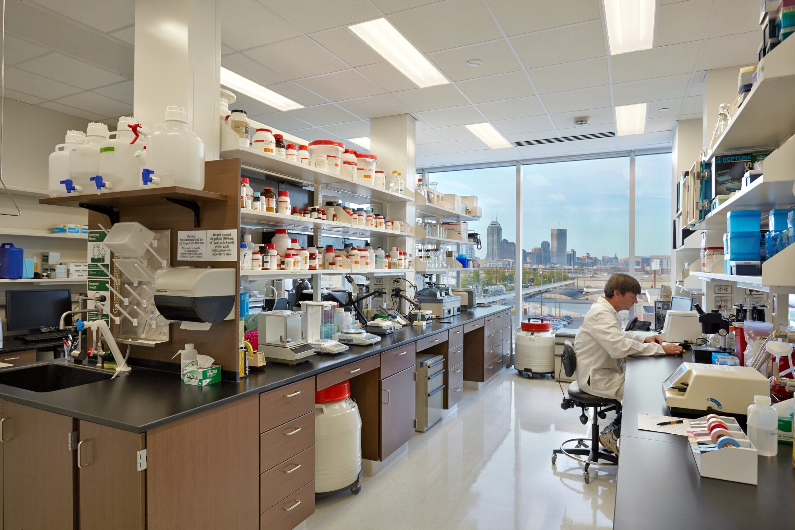 The lab is well-lit, with clean black countertops, multiple workstations including equipment like scales, microscopes, and other highly technical devices, and open shelving holding various lab equipment, glassware, and chemical containers. A student in a white lab coat is working on the right side on a computer. The floor is polished, and large windows in the background allow natural light to enter, showing a view of the Indianapolis skyline outside.