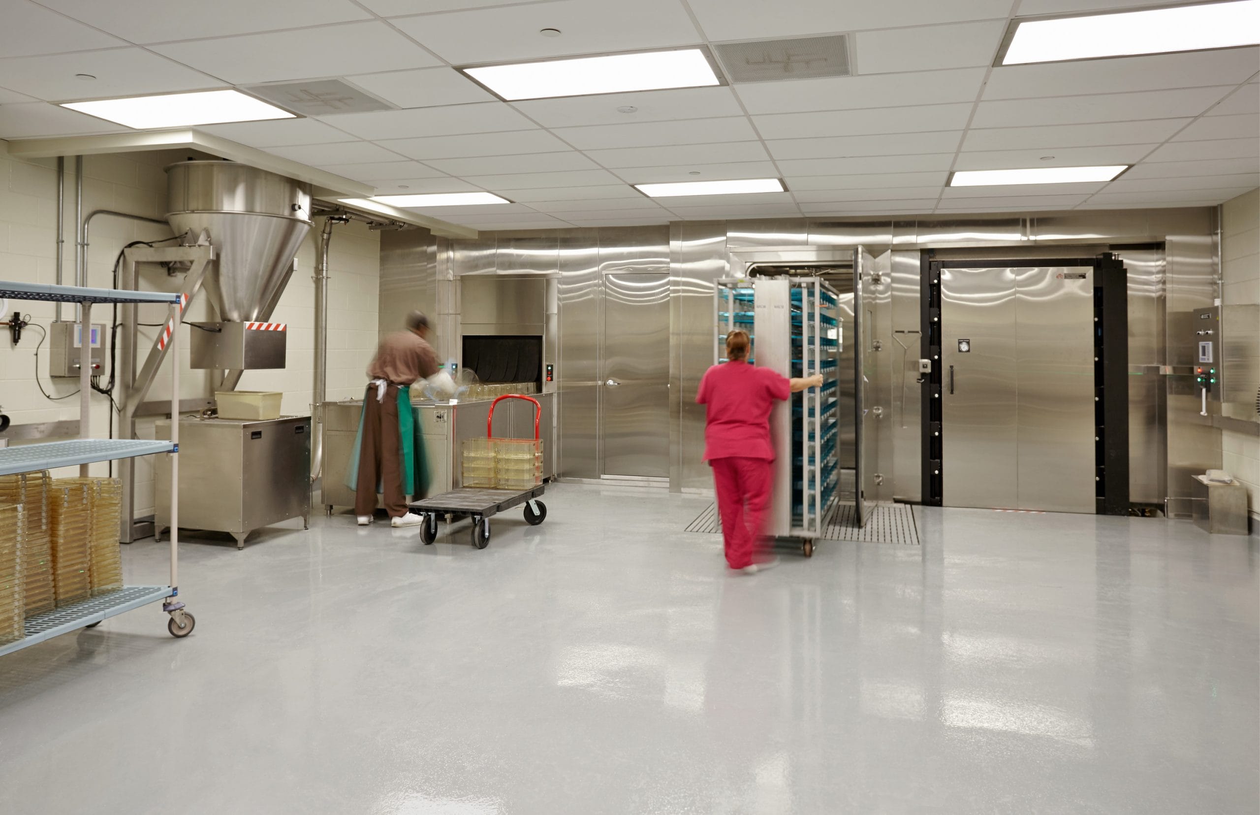A sterile laboratory or medical facility with stainless steel equipment and storage areas. Two workers are present: one wearing brown scrubs and an apron, handling lab trays near a large processing machine, while another in pink scrubs pushes a tall metal cart with multiple trays into a large industrial sterilizer or storage unit. The floor is highly reflective, and shelves stocked with petri dishes are visible on the left side of the room. The environment is brightly lit, with an emphasis on cleanliness and organization.