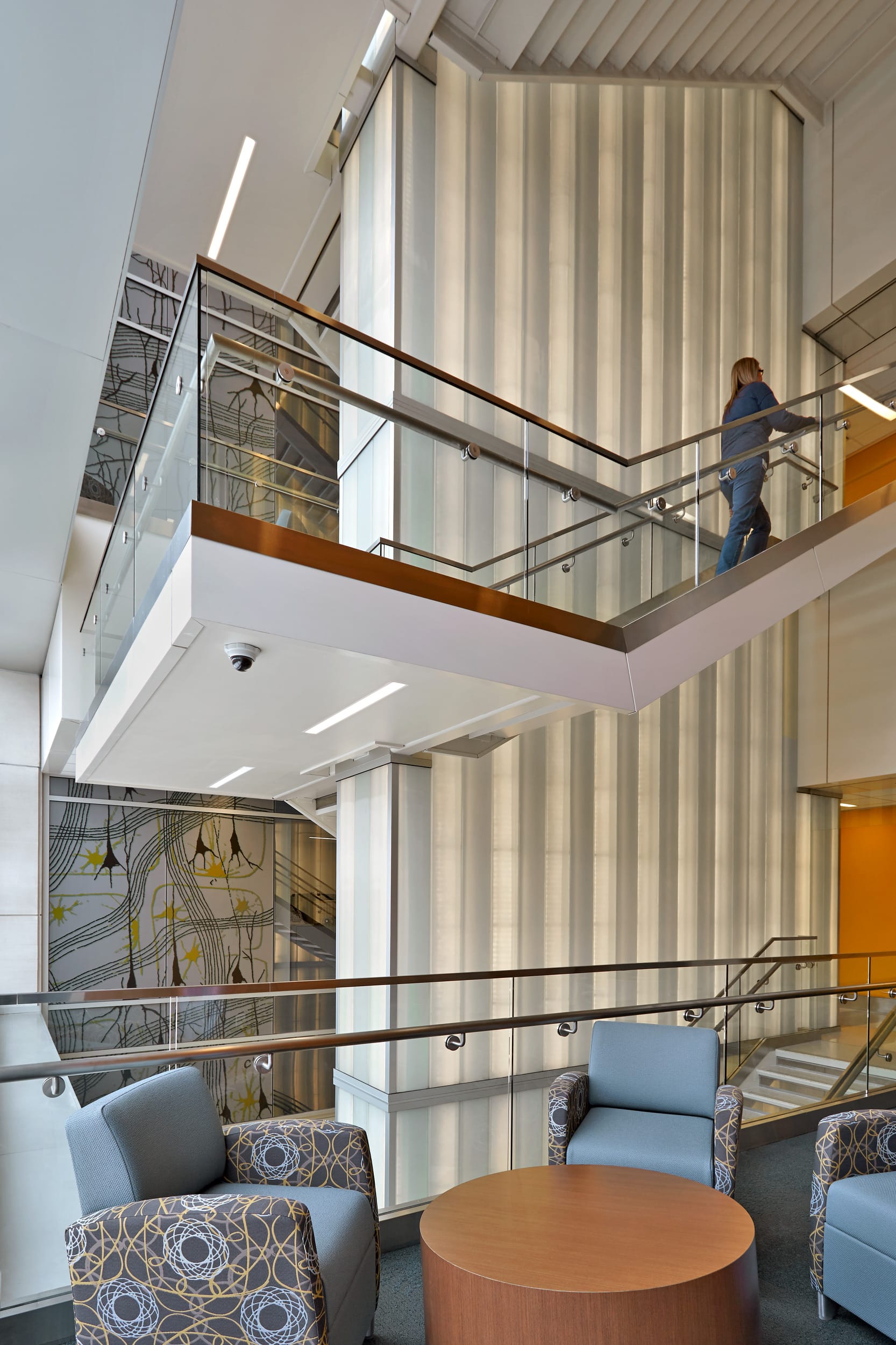 This image shows the interior of a modern building with a two-story open design. The space features a second-floor walkway with glass and metal railings with wooden handrails with person walking along the upper level. There are also light blue upholstered chairs and patterned accent chairs arranged around a round wooden coffee table in a seating area on the lower level. A tall vertical wall with ribbed or corrugated white paneling spans both floors and is lit from the inside. Behind it, a decorative wall panel or art installation with curved lines and yellow star-like elements is visible on the left side. The overall aesthetic is modern and bright.