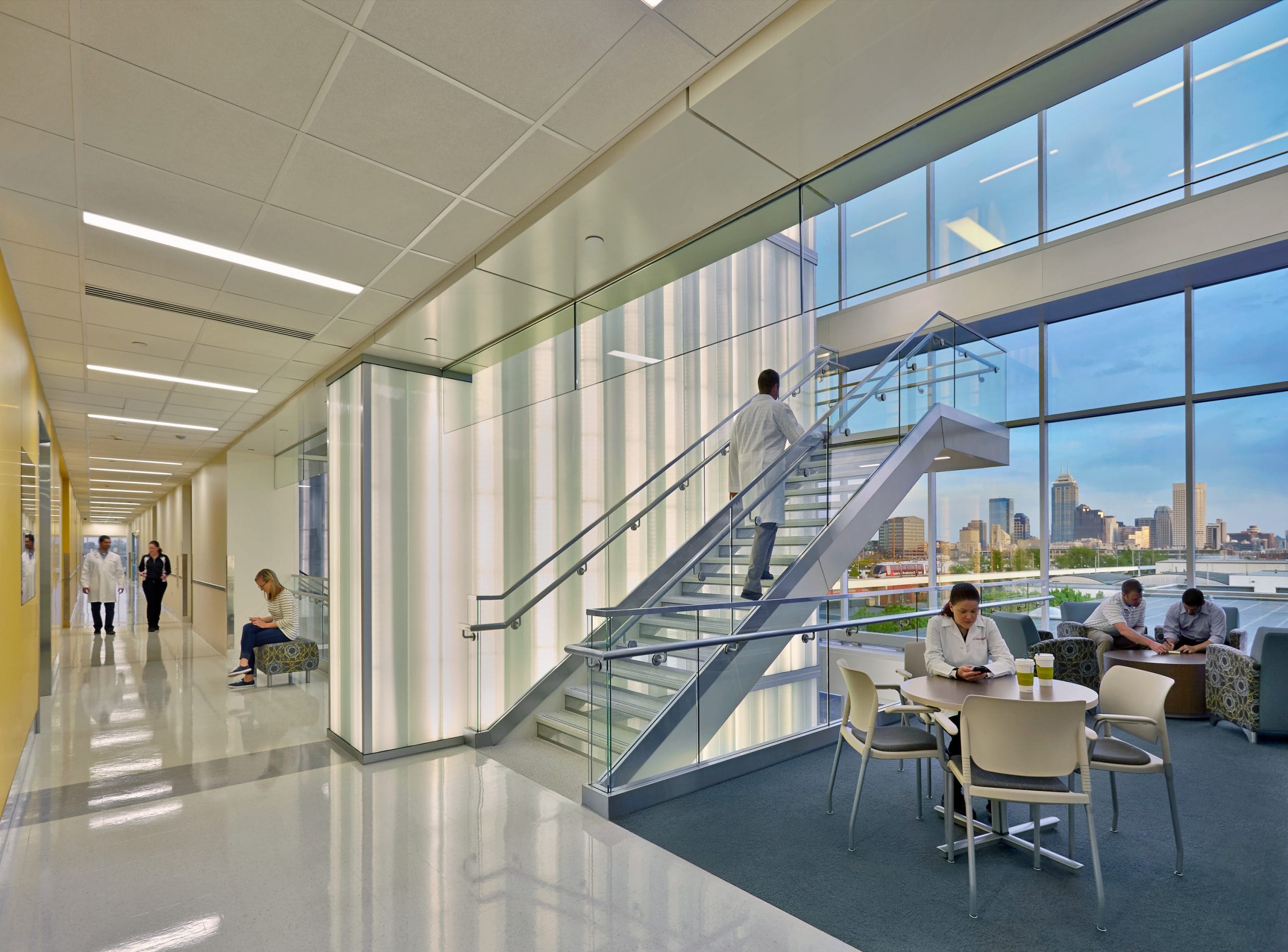 This image shows the interior of a modern building with a two-story open design. The space features a stairway with glass and metal railings with one person in a white lab coat walking along the upper level. Below the stairs is a seating area where another woman in a lab coat is enjoying a cup of coffee. A tall vertical wall with ribbed or corrugated white paneling stands alongside the stairs and is lit from the inside. Dramatic floor-to-ceiling multi-story windows allow for natural light and show a view of the Indianapolis skyline. The overall aesthetic is modern and bright.