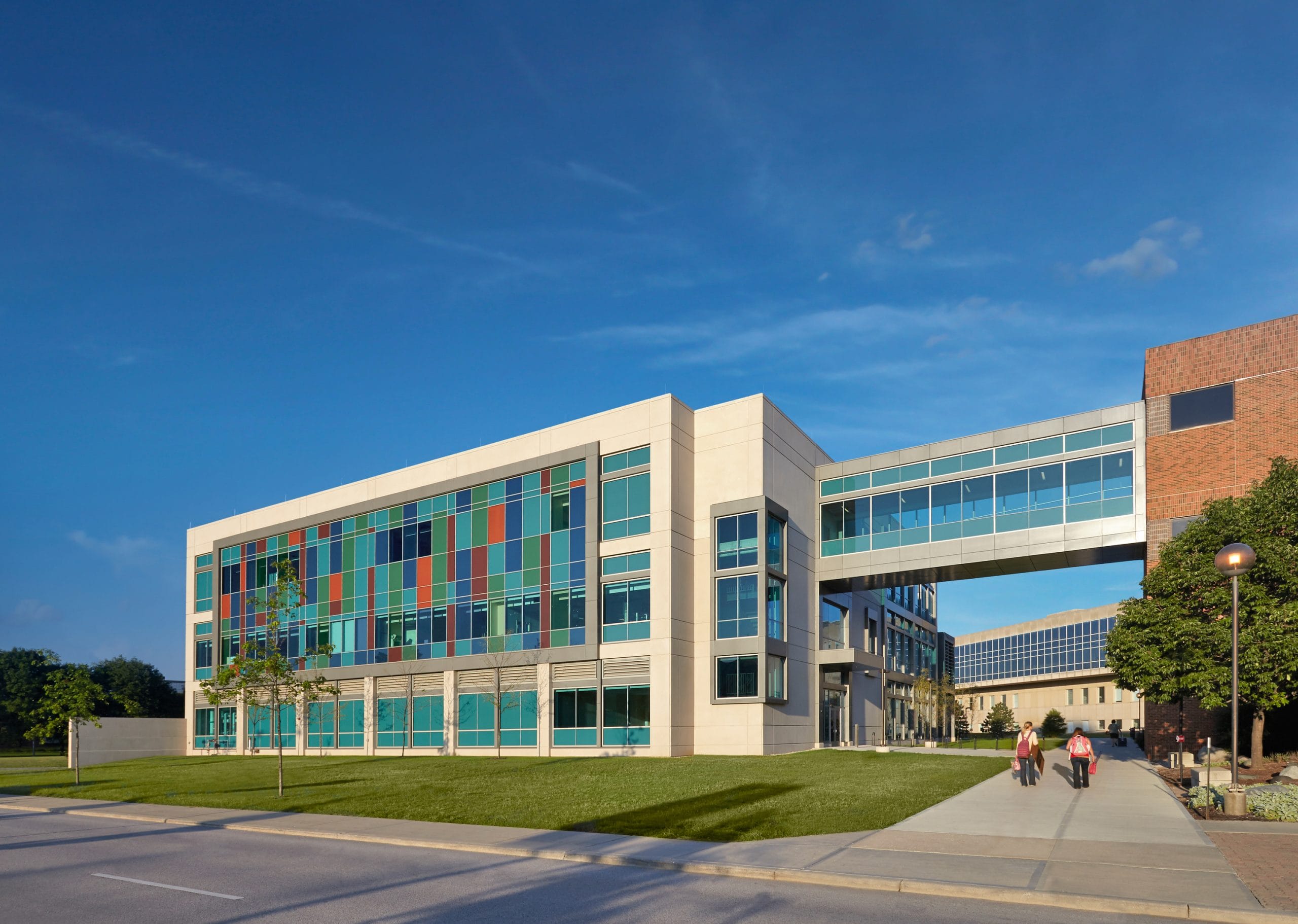 This image shows a modern building (the IU Science and Engineering Lab Building( featuring a striking architectural design with colorful stained glass or colored panel sections along the left façade. The structure has a white exterior with extensive glass windows and includes an elevated walkway or skybridge connecting to another building on the right. A few people can be seen walking on a concrete path in front of the building. The scene is captured on a bright sunny day with a clear blue sky, highlighting the vibrant colors of the glass panels which create a mosaic-like pattern. The building is surrounded by neatly maintained green lawns and some trees, with other campus structures visible in the background.