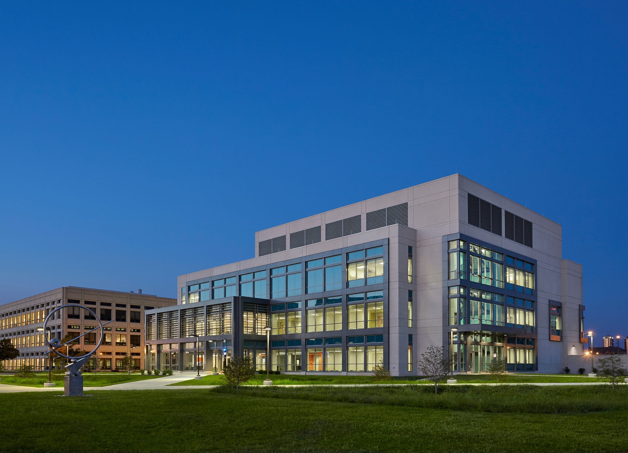 The image shows a modern institutional building (the Indiana University Science and Engineering Lab Building) photographed at dusk or early evening. The structure has a contemporary architectural design with a white exterior and extensive glass windows that are illuminated from within, creating a warm glow against the deep blue sky. To the left is another building with a similar institutional design, and between them is a landscaped area with green lawns and what appears to be a sculptural element or public art installation. The scene is peaceful with no people visible, and the lighting suggests it's after business hours when the buildings are still operational but the campus is quiet. The clean lines and large windows of the main building suggest it was designed to maximize natural light during daytime hours.