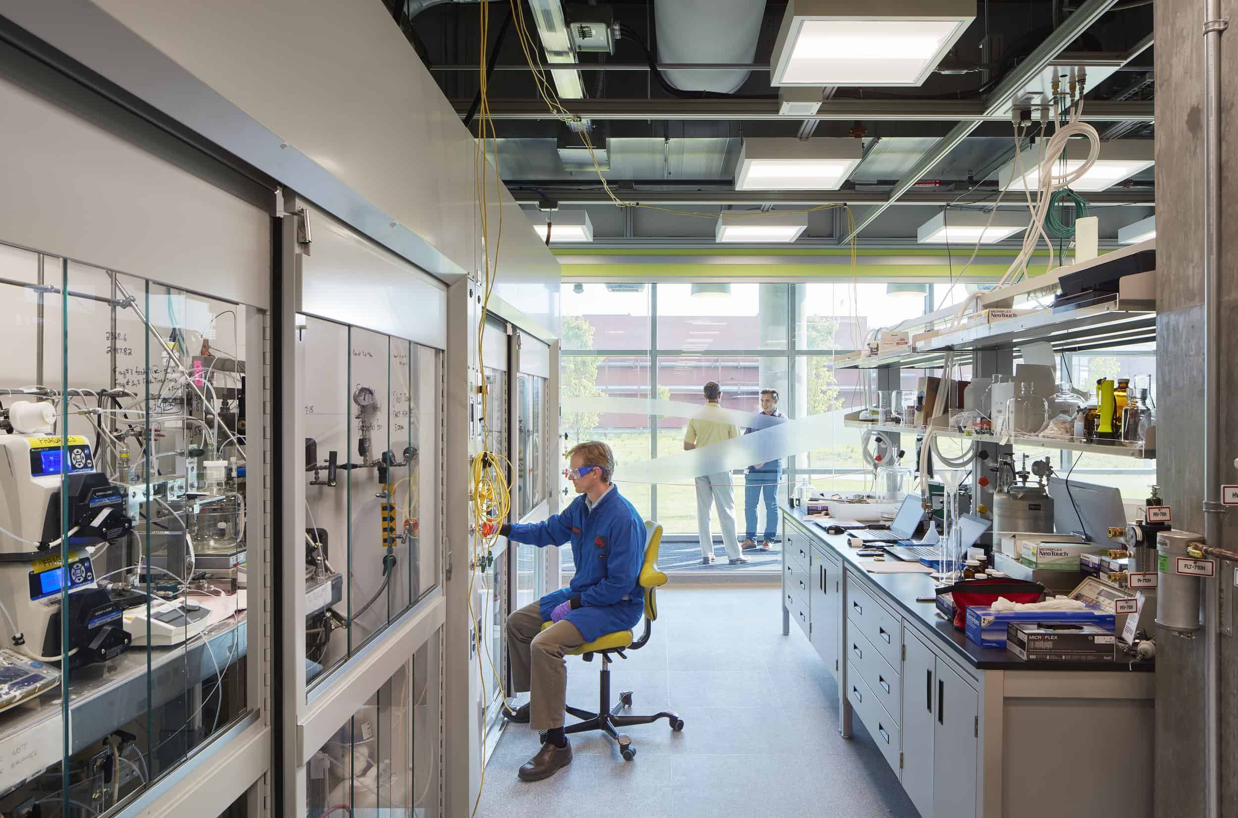 Modern research laboratory featuring a scientist in blue lab coat and safety glasses working at equipment mounted in glass-fronted cabinets. The well-equipped space has workbenches lined with instruments and supplies along the right wall, an exposed ceiling with utility connections, and large windows at the far end where two other researchers are reviewing documents. The bright, organized environment combines scientific functionality with natural light, creating an optimal setting for technical research and experimentation.