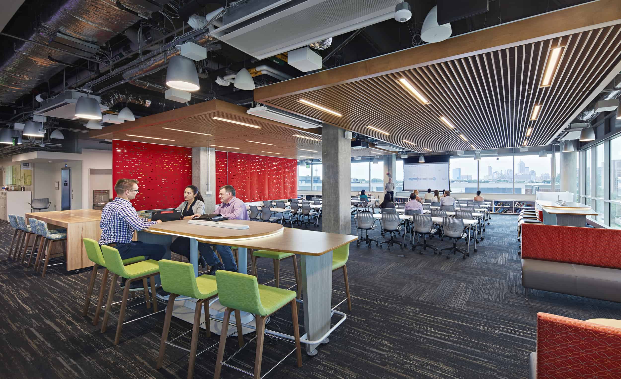 Modern open-concept office space featuring a blend of collaborative and individual workstations. In the foreground, three people converse at a wooden high-top table with bright green chairs. The space includes distinctive design elements like a striking red accent wall, wooden slat ceiling treatments, exposed industrial ceiling with pendant lighting, and large windows offering city views. The background shows additional workstations with ergonomic gray chairs, creating a contemporary workspace that balances aesthetics with functionality.