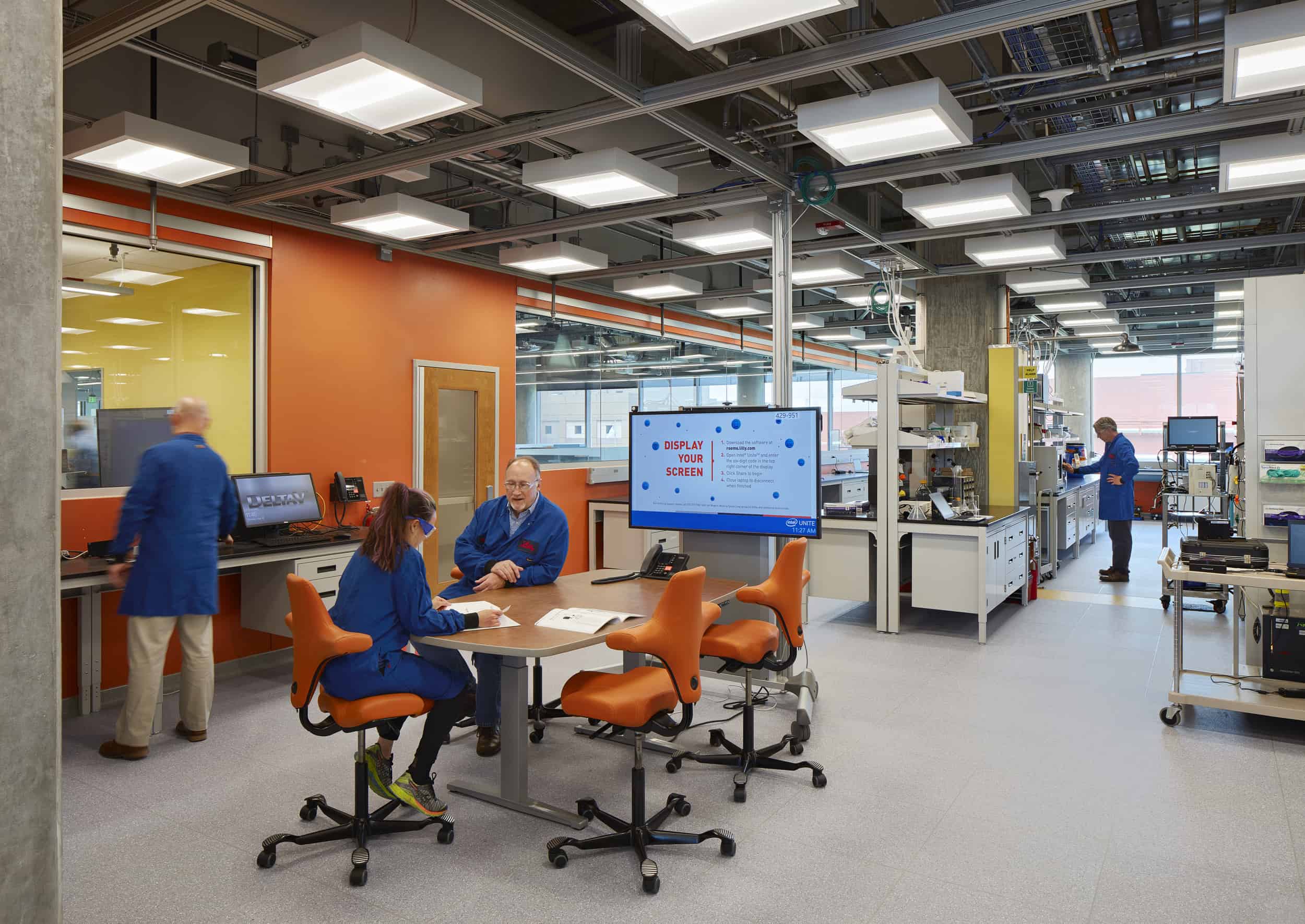 The image shows a modern laboratory or research workspace with vibrant orange walls and an industrial ceiling with exposed fixtures. In the foreground, two people wearing blue lab coats are seated at a round table with orange ergonomic chairs, reviewing documents together. Another person in a blue lab coat stands at a workstation to the left. The space features multiple workbenches with scientific equipment, computer monitors, and a display screen showing