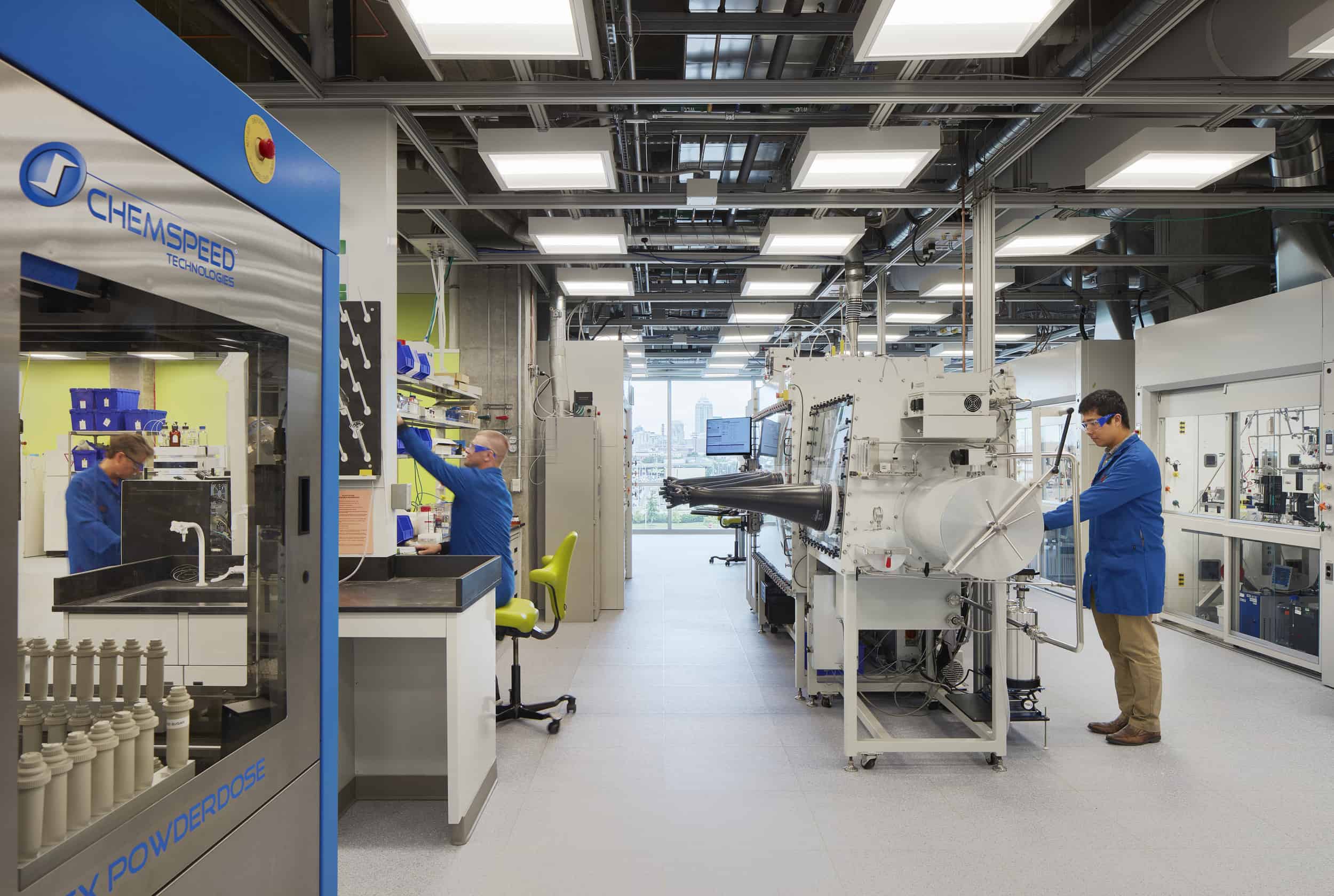 High-tech chemical engineering laboratory featuring three researchers in blue lab coats and safety glasses working with specialized equipment. The space includes advanced analytical instruments, including what appears to be a glove box system with black handling arms in the center and a