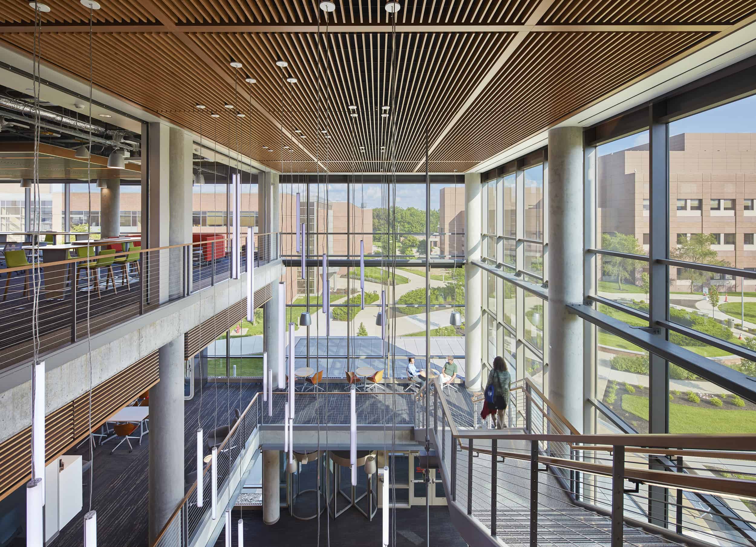 Modern multi-story academic or research building interior featuring a dramatic atrium with floor-to-ceiling windows showcasing a landscaped campus view. The space incorporates sustainable design elements including wooden slat ceilings, exposed concrete columns, and cable railings along multiple levels. Natural light floods the open space, illuminating seating areas on different floors. The upper level shows collaborative spaces with colorful furniture, while a few people can be seen on the lower level near the glass façade. This contemporary architectural design creates visual connections between interior spaces while maintaining a strong relationship with the surrounding campus environment.