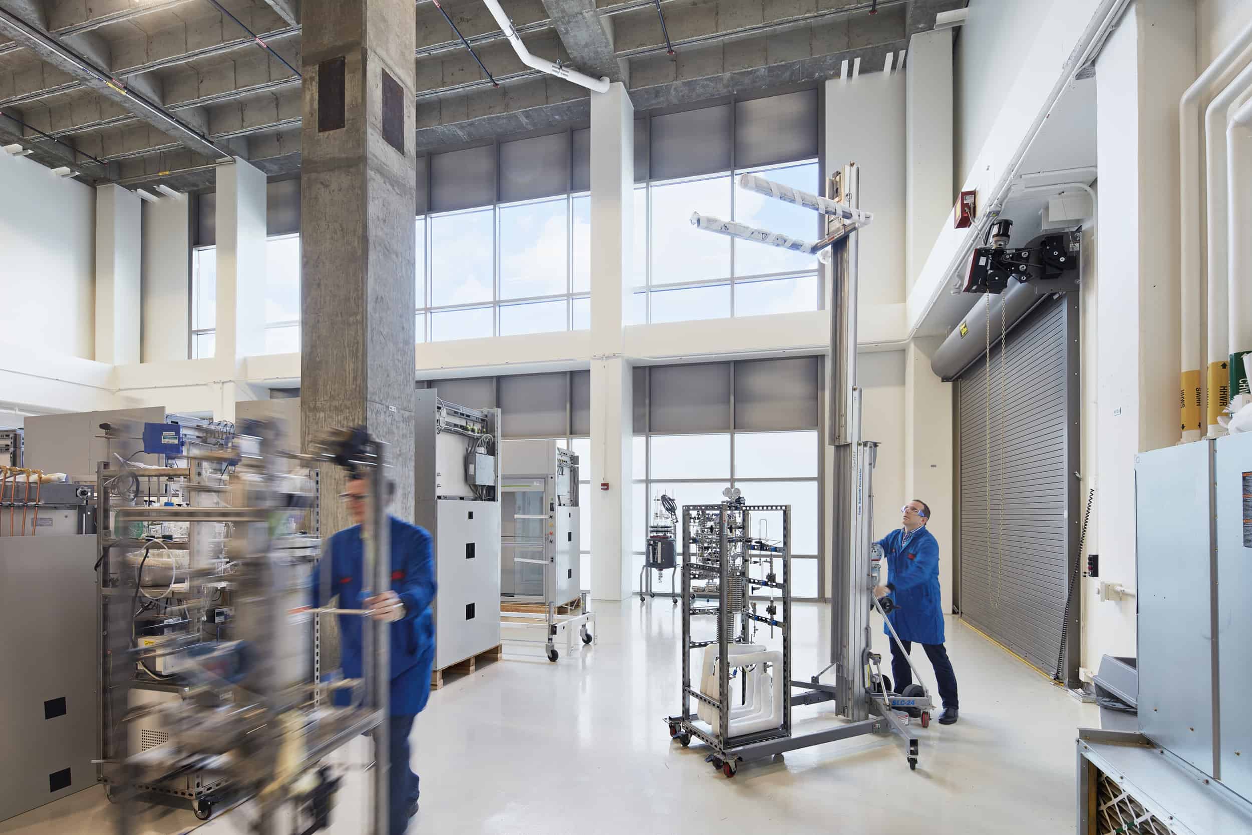 Spacious industrial research facility with high ceilings and abundant natural light from floor-to-ceiling windows. Two researchers in blue lab coats are working with specialized equipment - one pushing a cart (shown in motion blur on the left) and another operating mechanical apparatus on the right. The space features exposed concrete columns and ceiling infrastructure, polished white floors, and various scientific instruments including what appears to be complex testing equipment on movable racks. The modern laboratory design combines functional workflow areas with architectural elements that create an open, light-filled environment for technical research work.