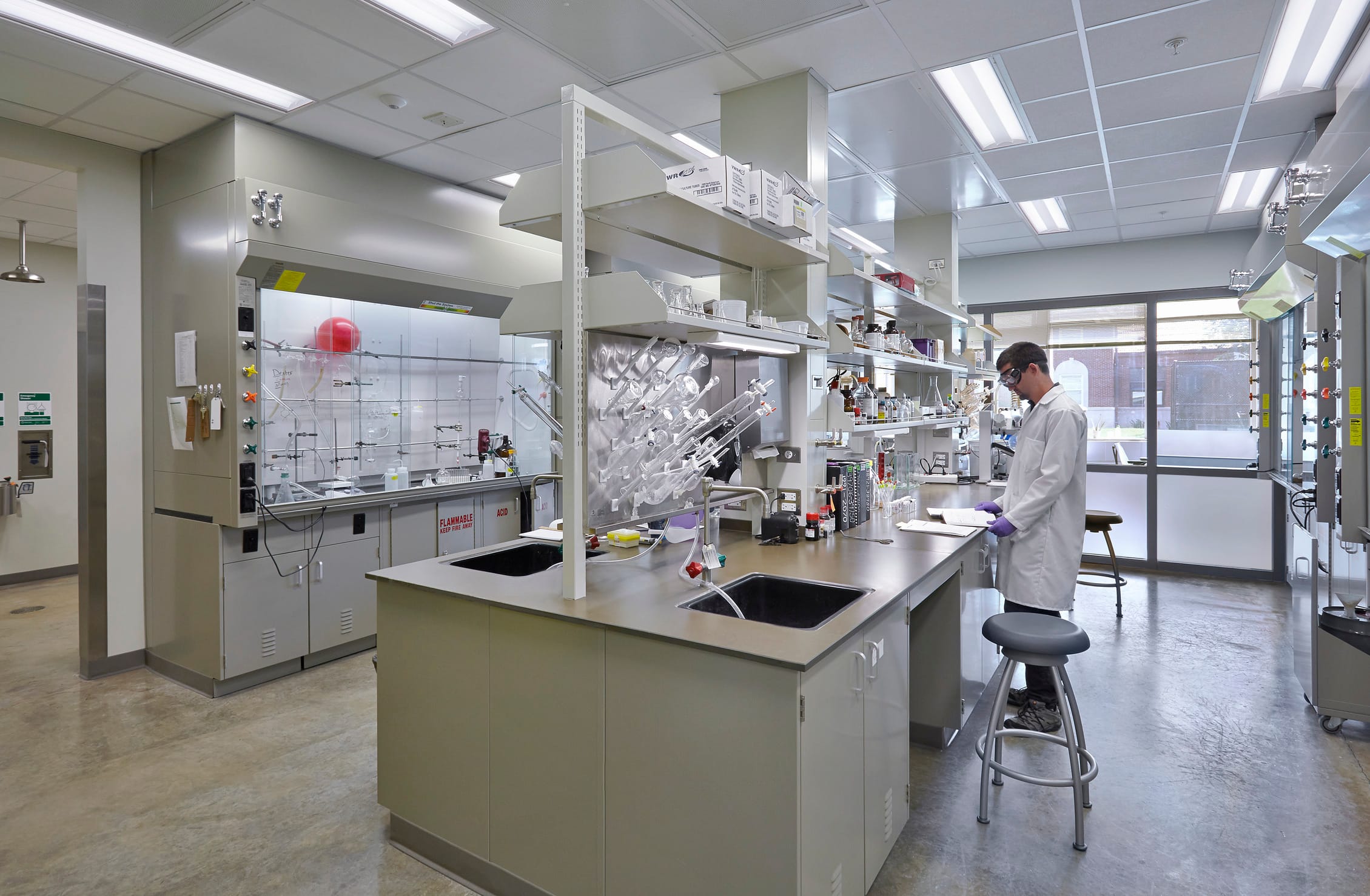 A modern laboratory with a researcher in a white lab coat, safety goggles, and purple gloves standing at a workstation, writing on a clipboard. The lab is well-lit, with stainless steel countertops, multiple sinks, and open shelving holding various lab equipment, glassware, and chemical containers. A fume hood with a glass front and labeled controls is visible on the left, along with a drying rack for glassware. The floor is polished concrete, and large windows in the background allow natural light to enter, showing a view of a building outside.
