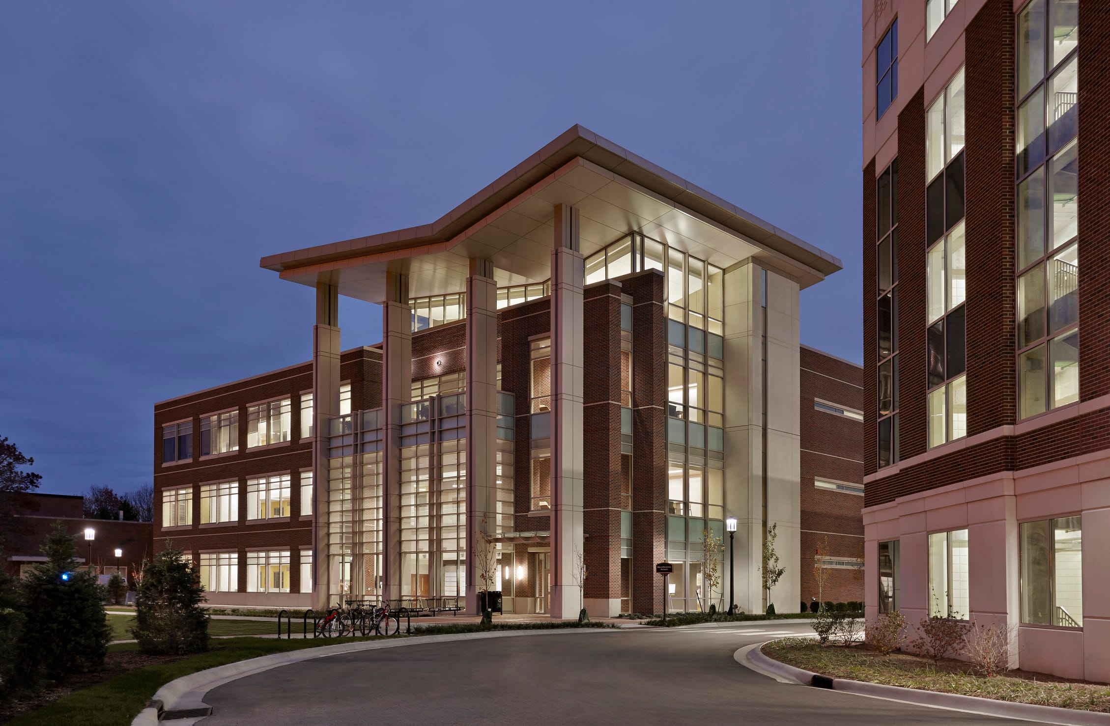 A modern academic building captured at dusk with lights from within the 4-story building illuminating its architectural features. The structure combines brick, glass, and metal elements, with tall, rectangular columns supporting an extended roof. Sleek metal accents and horizontal louvers add to the contemporary design.