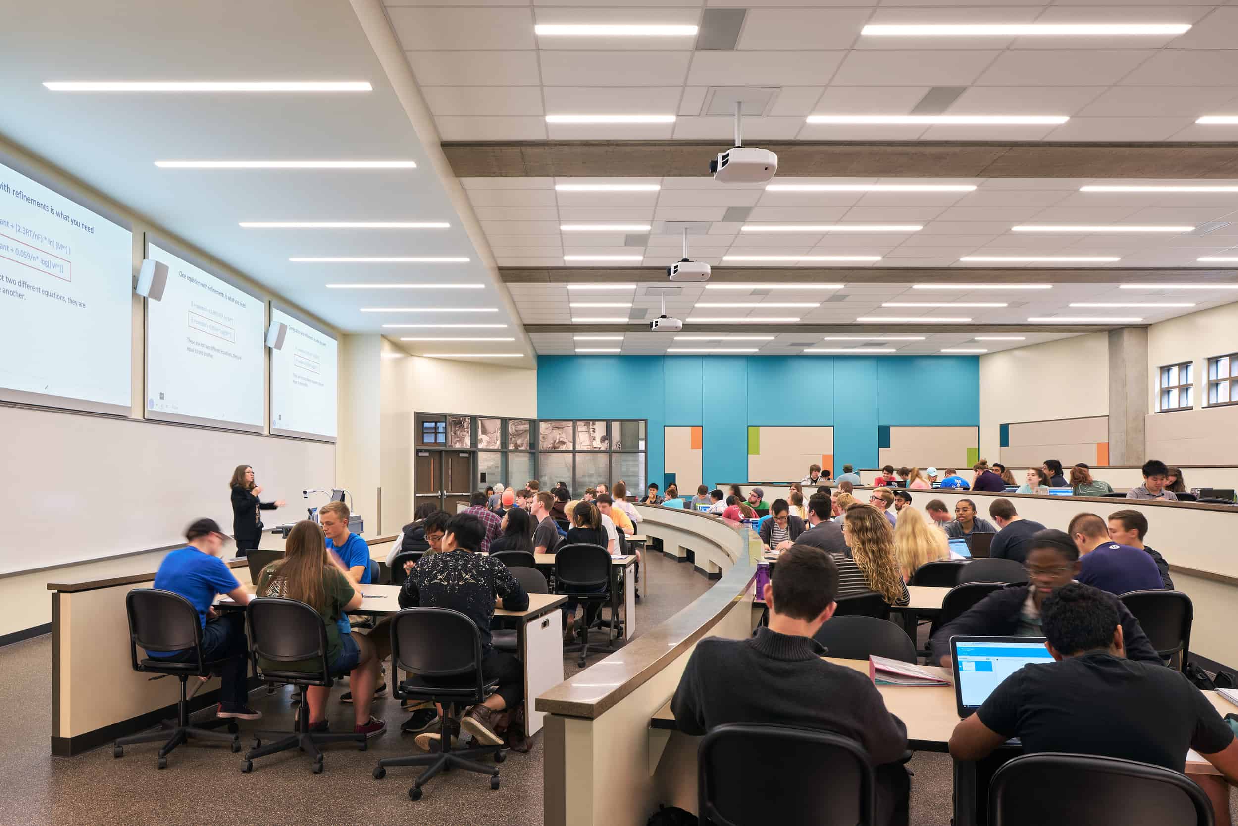 This image shows a modern university lecture hall with a tiered, semi-circular seating arrangement. Students are seated at curved desks with black office chairs, many working on laptops or in small groups. The room features multiple projection screens on the left wall displaying presentation slides with text that's not clearly legible in the image. At the front of the room, an instructor is standing near the projector screens. The classroom has a contemporary design with bright fluorescent lighting in the drop ceiling, multiple ceiling-mounted projectors, and a distinctive teal blue accent wall at the back of the room with some display panels. Small windows near the ceiling allow for some natural light. The space appears designed for collaborative learning, with students arranged in a way that allows for both viewing the presentation and working together in small groups.