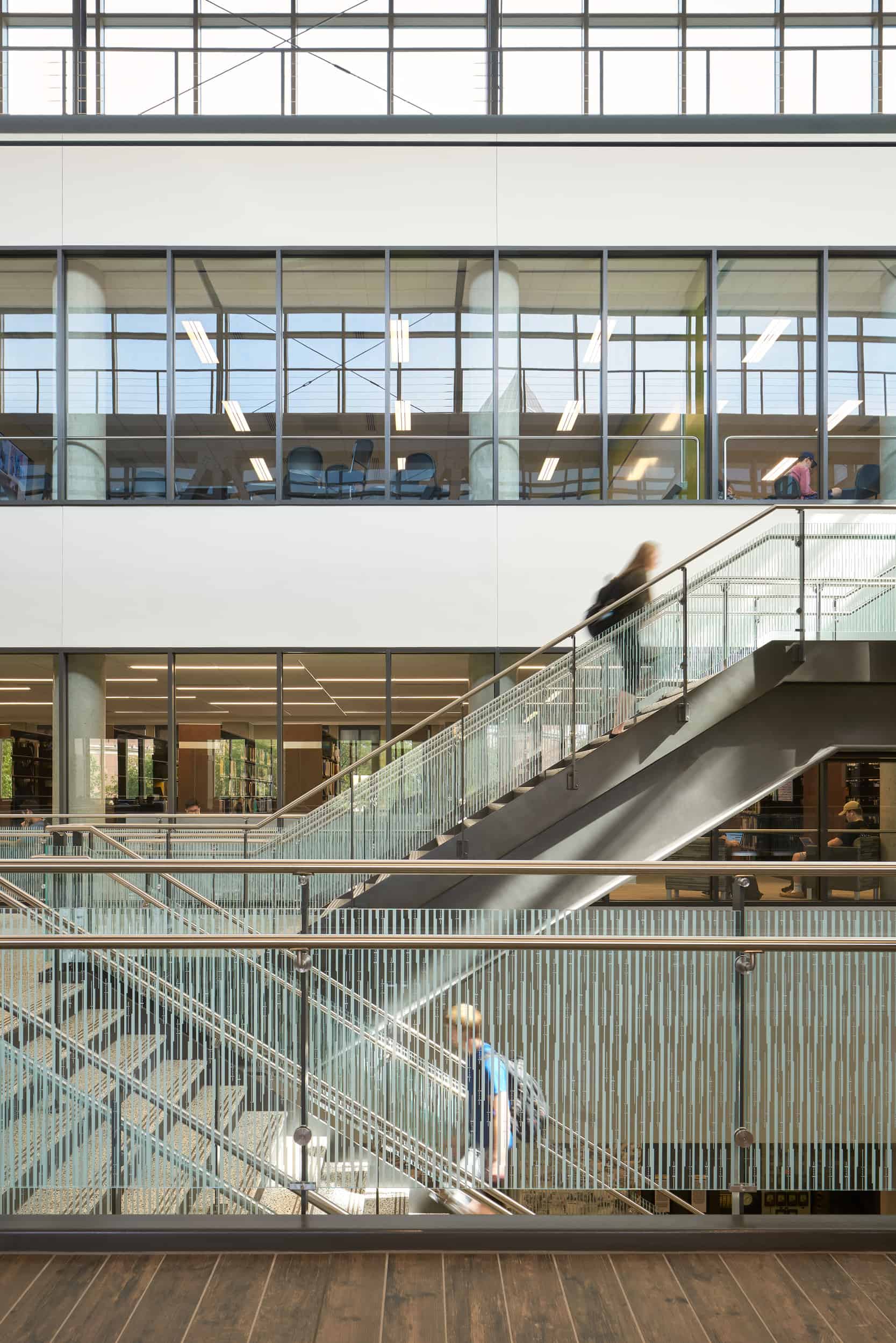 A spacious multi-level atrium in the Active Learning Center at Purdue University. The central feature is a large staircase with glass railings leading to multiple floors, with people ascending and descending. Three floors of classrooms and workspaces are visible beyond the staircase through floor-to-ceiling interior glass walls. The walkways and staircases create a contemporary industrial aesthetic, complemented by concrete columns and glass partitions.