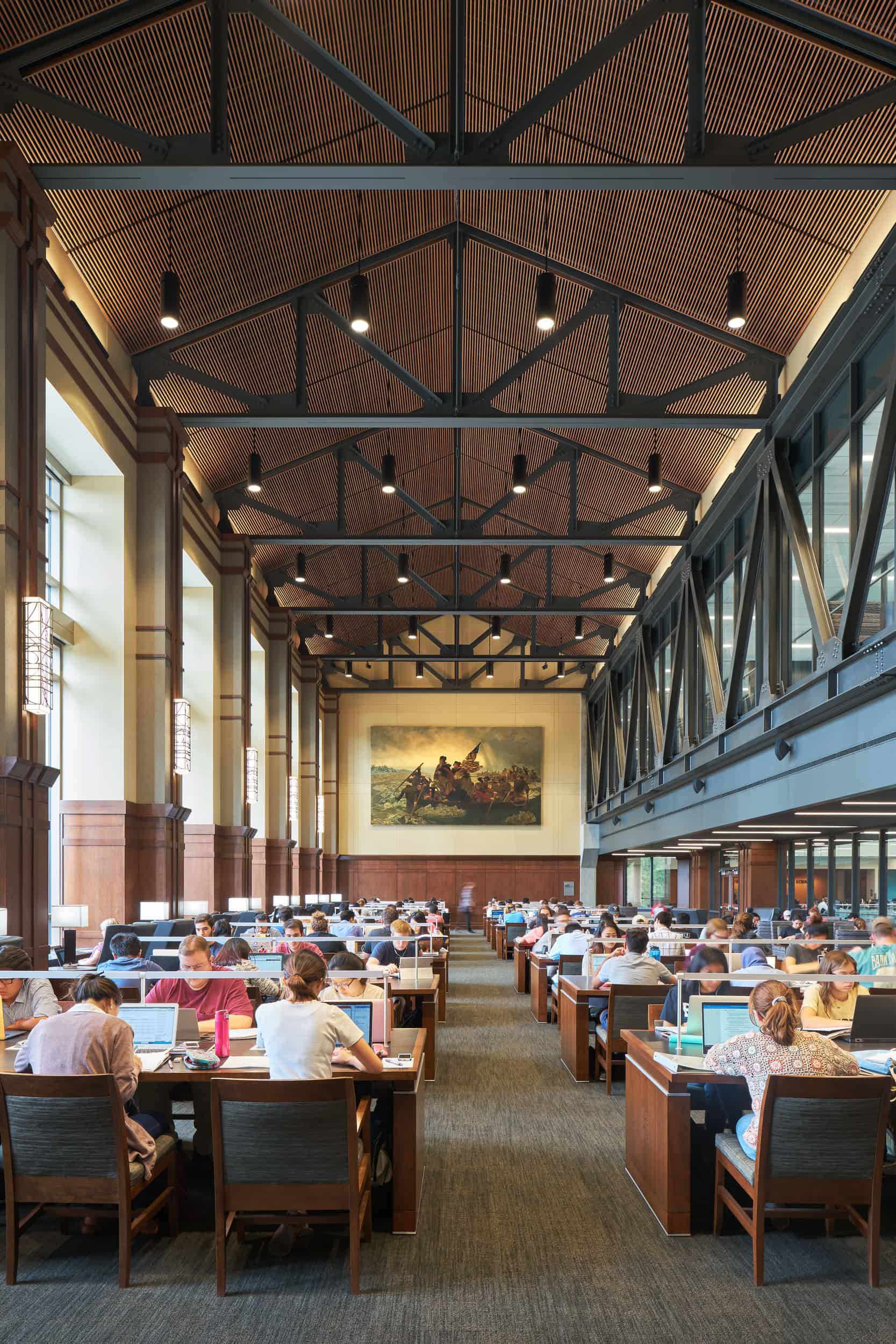This image shows a spacious university study hall at Purdue University. The interior features a grand, high ceiling with exposed wooden truss beams and an elegant slatted wood ceiling treatment. Pendant lights hang from the ceiling, providing warm illumination throughout the space. The room is filled with wooden study tables where numerous students are working at laptops or studying. The hall has a traditional academic aesthetic with wood paneling along the lower walls and large windows allowing natural light to enter. At the far end of the hall, a large historic framed painting is prominently displayed on the wall. The space appears to be a blend of historic architectural elements with modern functionality, creating an impressive and conducive environment for studying.