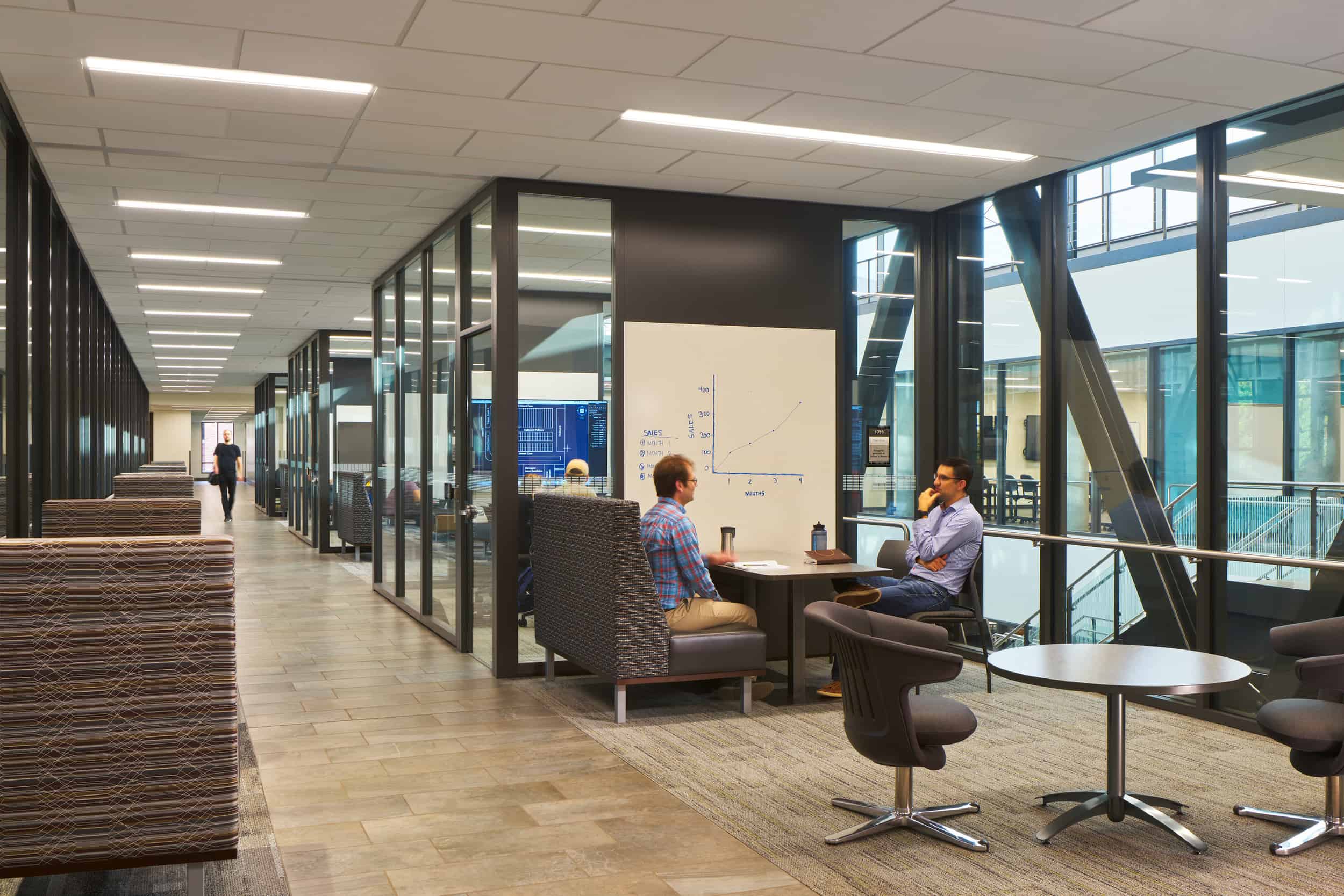 This image shows a modern educational collaborative space featuring semi-private meeting areas with glass walls. In the foreground, two people are seated in a small meeting pod engaged in discussion, with a whiteboard visible containing what appears to be a graph or chart. The seating area includes comfortable upholstered chairs around a small table. The space has an open, contemporary design with a long corridor that extends into the distance, where another person can be seen walking. The left side of the corridor has modular, striped seating elements or storage units. The right side features several glass-enclosed meeting rooms or collaboration spaces. The interior design incorporates natural light from large windows along the right side, modern recessed ceiling lighting, and a mixed-material floor with wood-look tiles in the corridor and carpeted areas in the meeting spaces. The overall aesthetic emphasizes transparency, collaboration, and a blend of open and semi-private workspaces typical of modern academic buildings or corporate innovation centers.