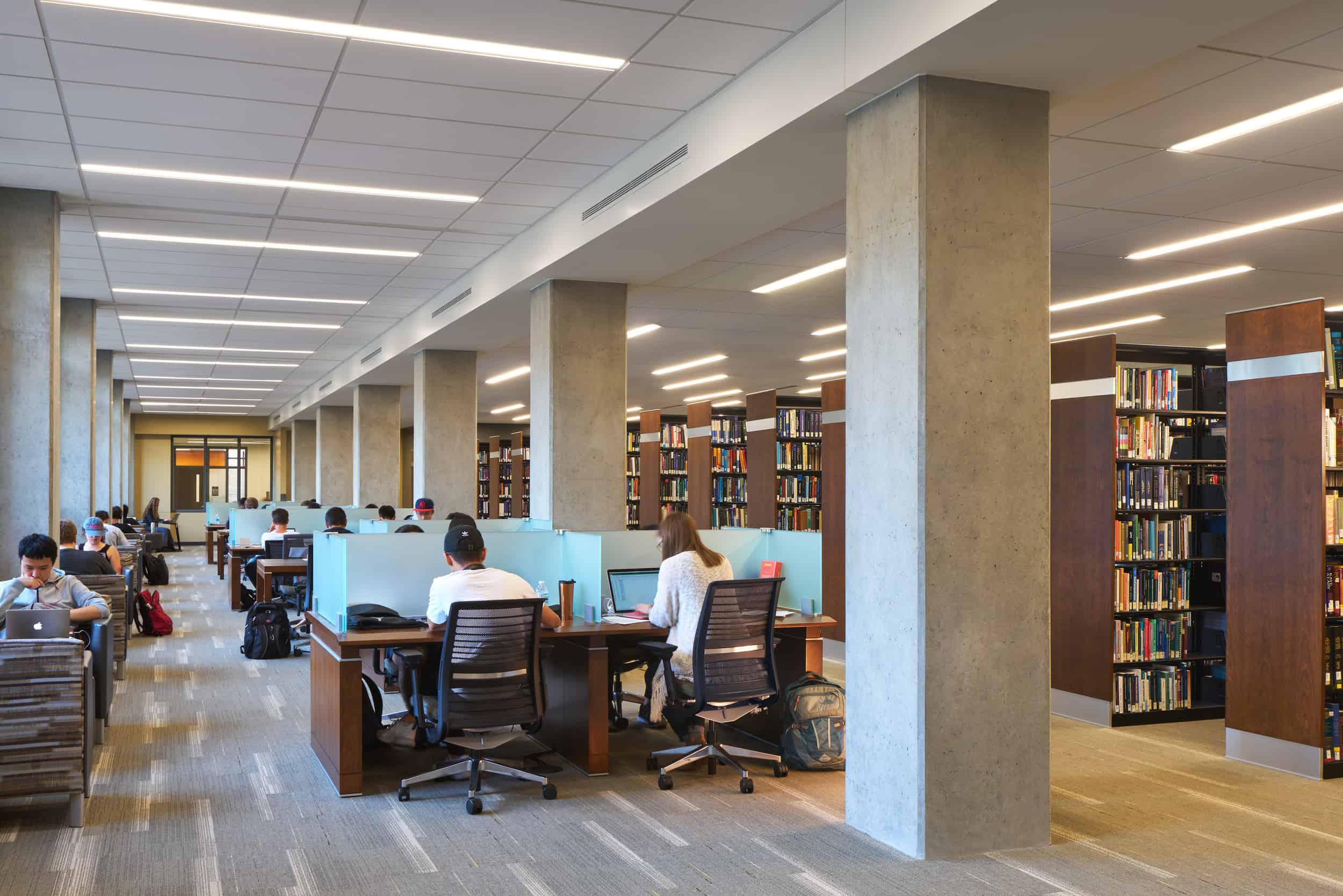A modern academic library in the Purdue University Active Learning center with rows of study carrels occupied by students working on laptops and reading. The space features a combination of individual study desks with frosted glass dividers and open seating areas. Tall concrete columns support the ceiling, which has a grid of recessed lighting. Wooden bookshelves filled with books line the right side of the image, while students are scattered throughout, focused on their work. Some students have backpacks and personal items at their desks. The atmosphere is quiet and studious, with a mix of natural and artificial lighting.