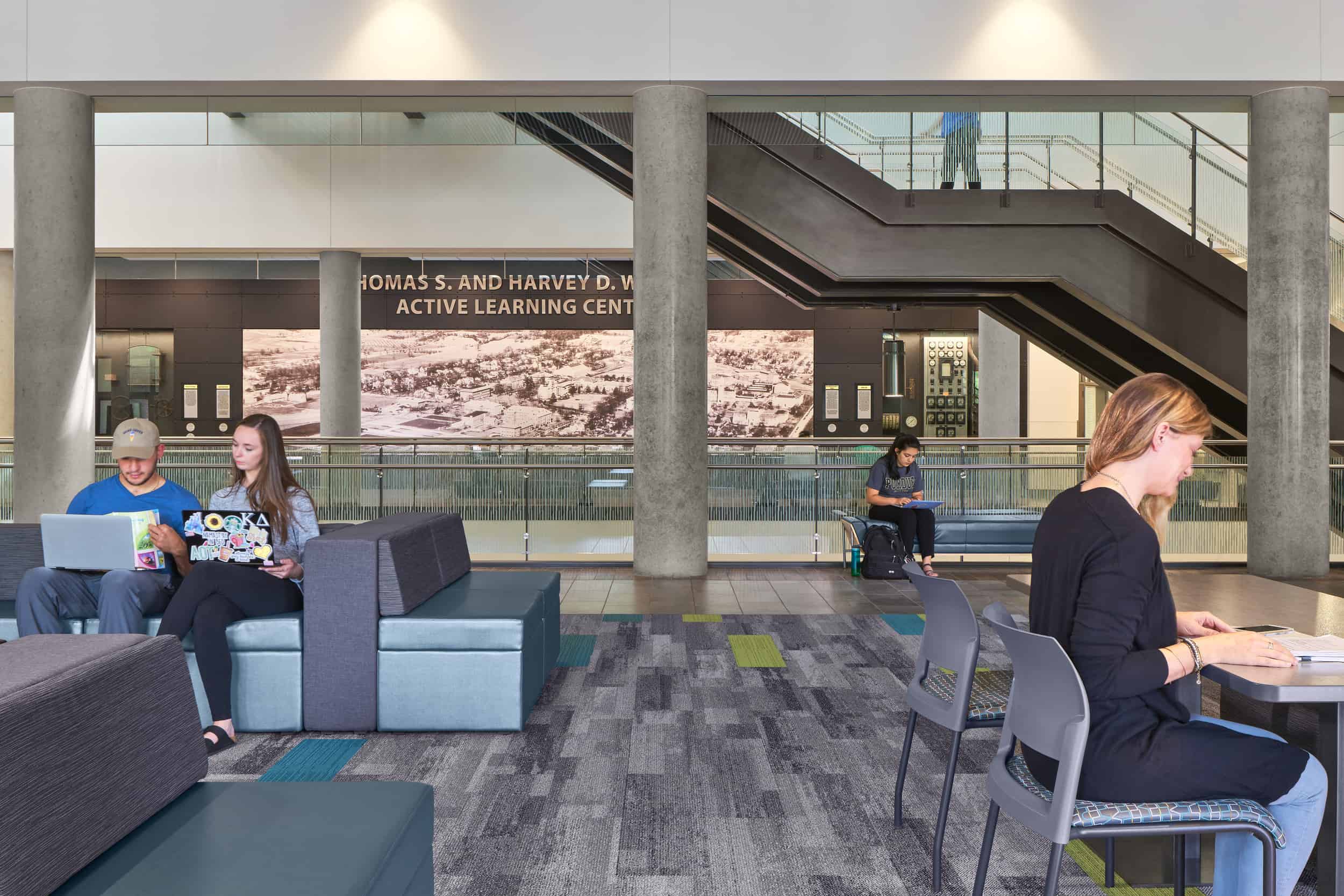 A modern, open-concept learning space within the Thomas S. and Harvey D. Wilmeth Active Learning Center. Several students are engaged in different activities—two students are collaborating on laptops while sitting on modular seating, another student is studying at a table, and one more is sitting near a railing, reading with a backpack beside them. The area features contemporary furniture, a mix of carpet and tile flooring, and large concrete columns. In the background, escalators lead to an upper level, and a historic aerial photograph decorates the wall, contributing to the academic atmosphere