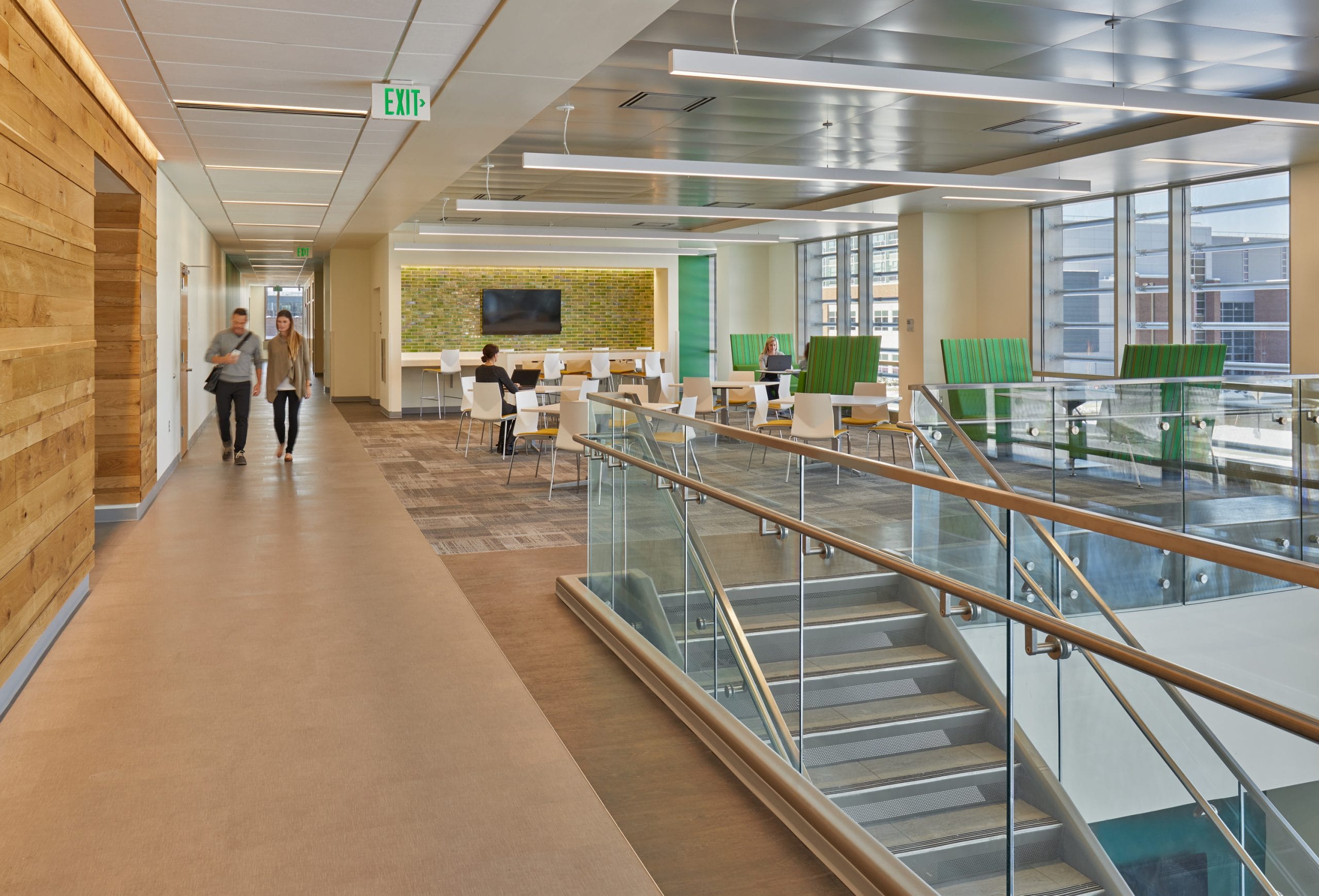 A view from the second floor of modern, multi-level lobby in the Hobart and Russell Creighton Hall of Animal Sciences featuring a striking glass and metal staircase coming up to a common area with bright green furniture. The bright, open space includes a seating area with vibrant green chairs around a white circular table. Natural light floods through floor-to-ceiling windows, complementing the wood-paneled accent walls and green tile wall with a television. The design combines contemporary architectural elements with comfortable collaborative spaces for students and faculty.
