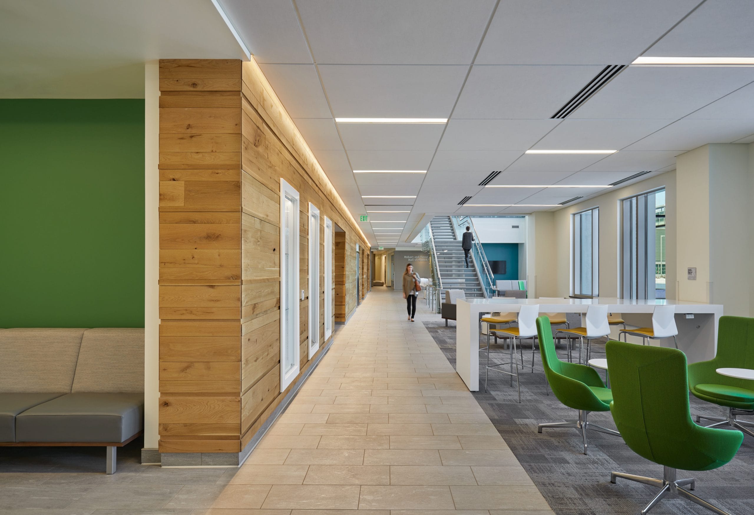 A view of the modern, multi-level lobby in the Hobart and Russell Creighton Hall of Animal Sciences from deeper inside the building. From this view, the striking glass and metal staircase in the atrium with a person ascending is still visible. The bright, open space includes a seating area with vibrant green chairs on grey carpet, distinguished from a tile walkway. There are wood-paneled accent walls that tie in the agricultural studies that happen within the building. The design combines contemporary architectural elements with comfortable collaborative spaces for students and faculty.